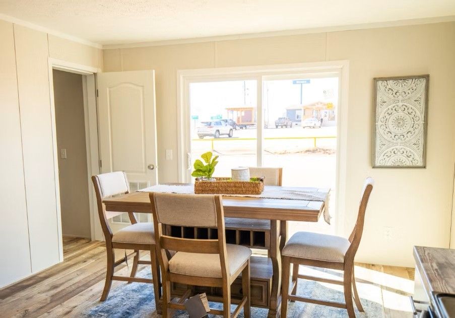 Dining area with wooden table and chairs, sunlight through a large window, decorative wall art.