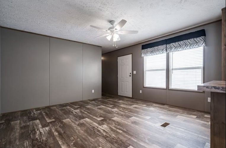 Empty living room with wood-look flooring, gray walls, ceiling fan, and window with blinds.