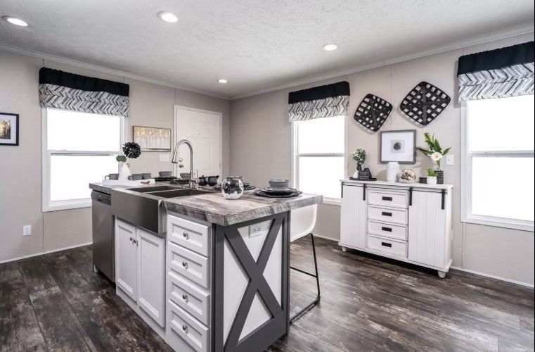 Kitchen with island, stainless steel sink, white cabinets, grey countertop, dark flooring, and window treatments.