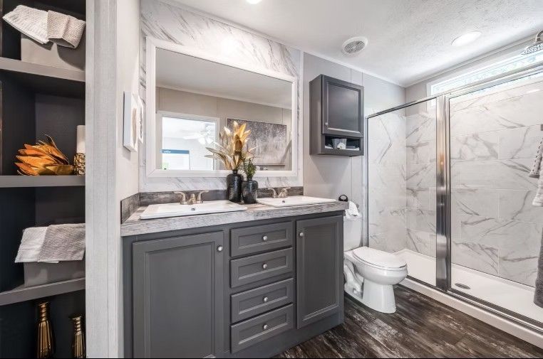 Bathroom with double sink vanity, shower, and toilet. Gray cabinets, marble-look walls, and dark wood-look floor.