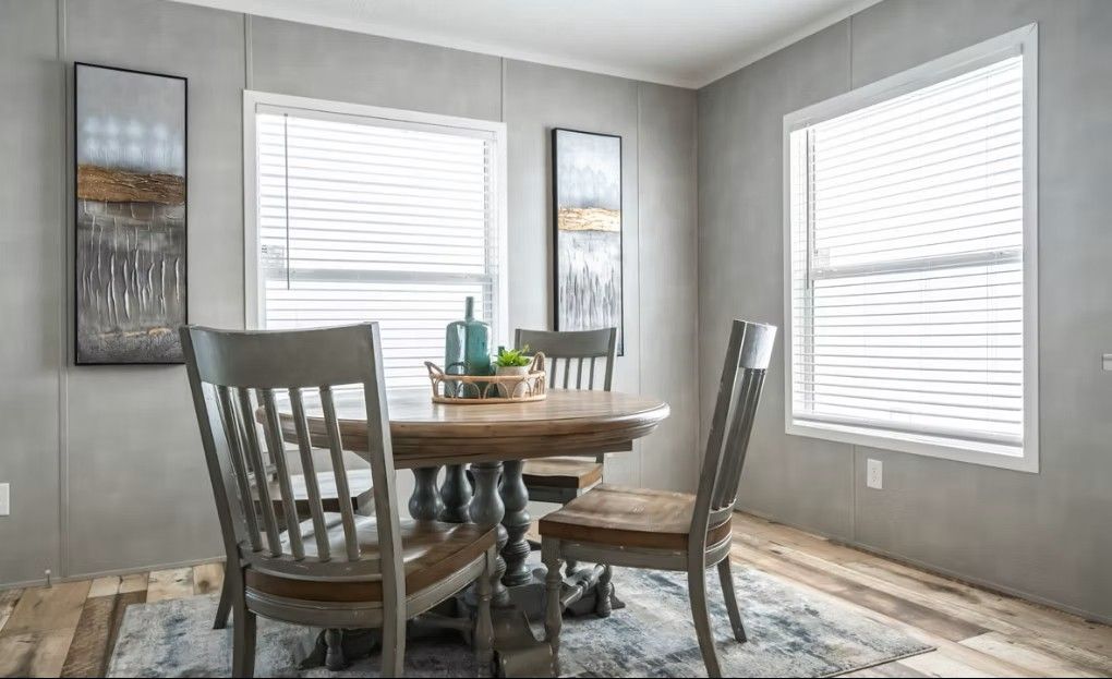 Dining room with round wooden table, chairs, art, windows with blinds, and rug.