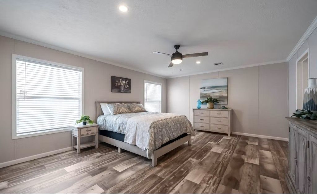 Bedroom with bed, dresser, nightstand, and two windows. Wood-look flooring and light gray walls.