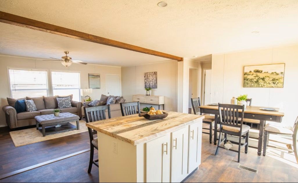 Open-plan living space: kitchen island, dining table, sofa, and artwork. Warm sunlight, wood beams, and neutral tones.