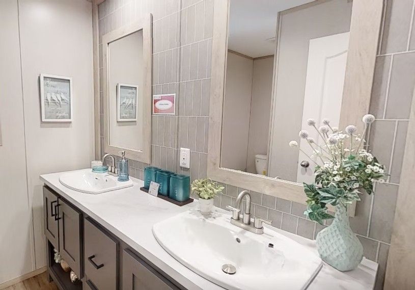 Bathroom with double sinks, gray cabinets, light gray tile wall, large mirrors, and decorative vase.