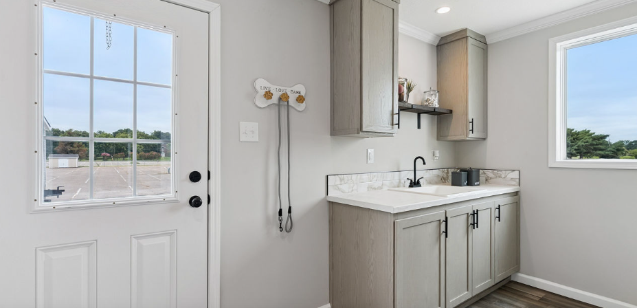 Bright laundry room with white door, utility sink, and gray cabinets next to a window