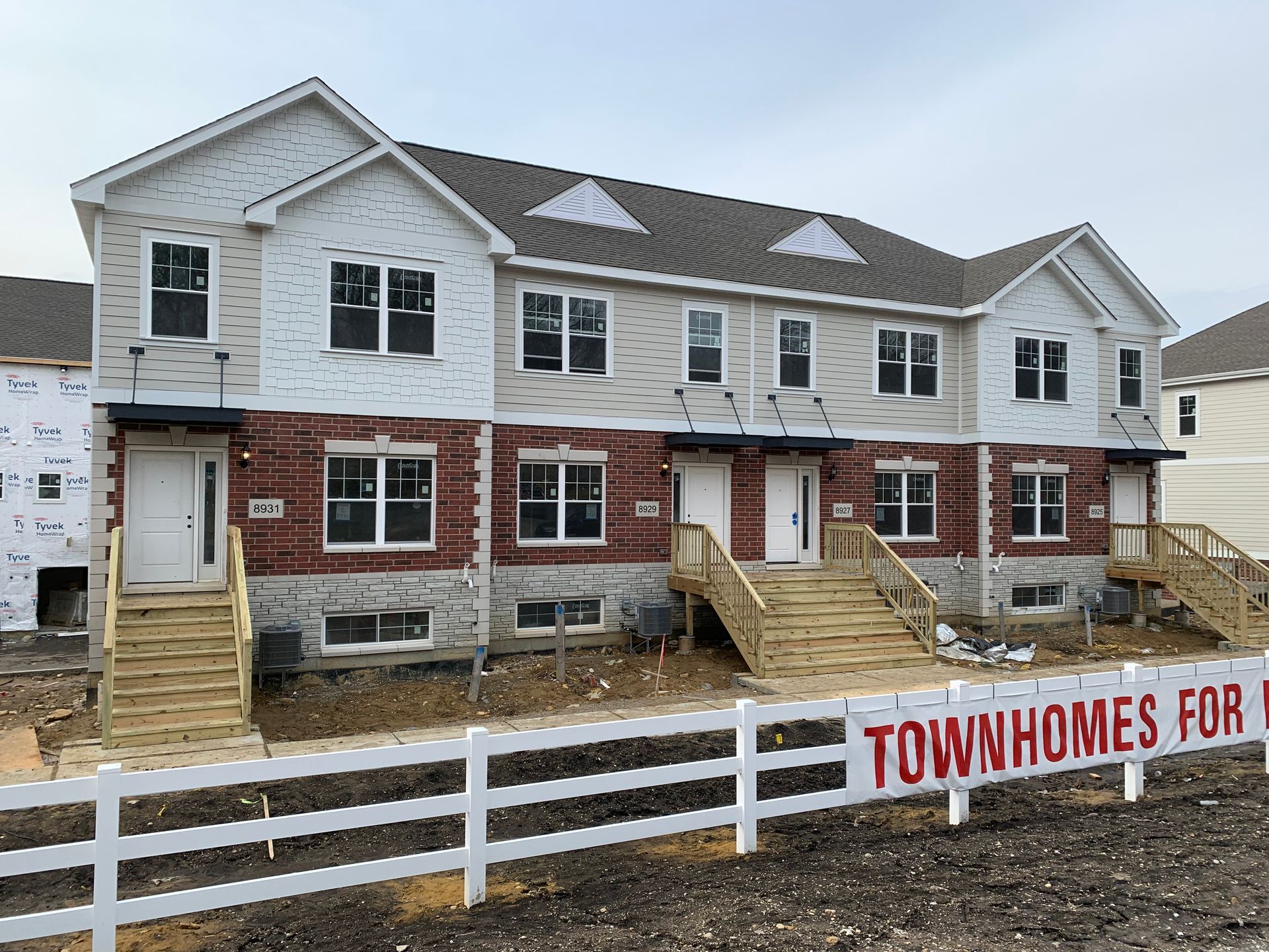 Townhomes under construction with gray siding, brick fronts, front steps, and a fence sign reading “TOWNHOMES.”