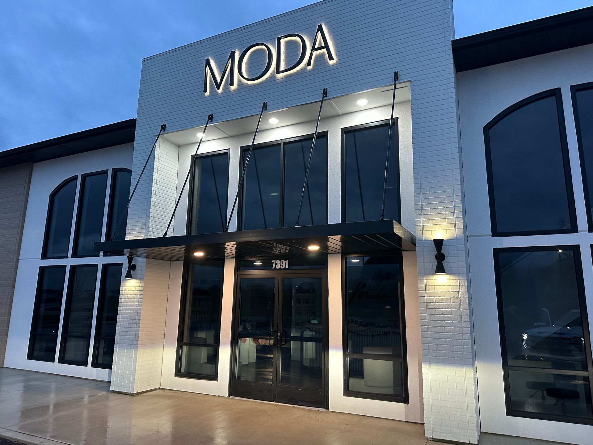 MODA storefront at dusk with glass doors, lit entrance, and blue sky overhead