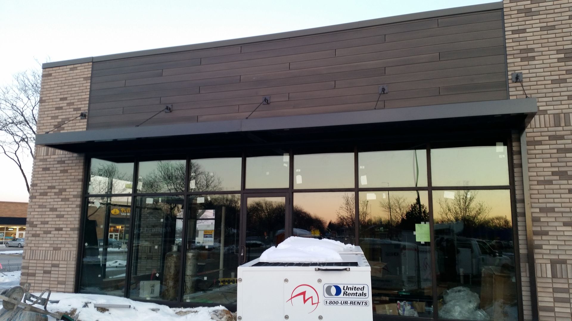 Storefront with large glass windows and a snow-covered roof, viewed at dusk.