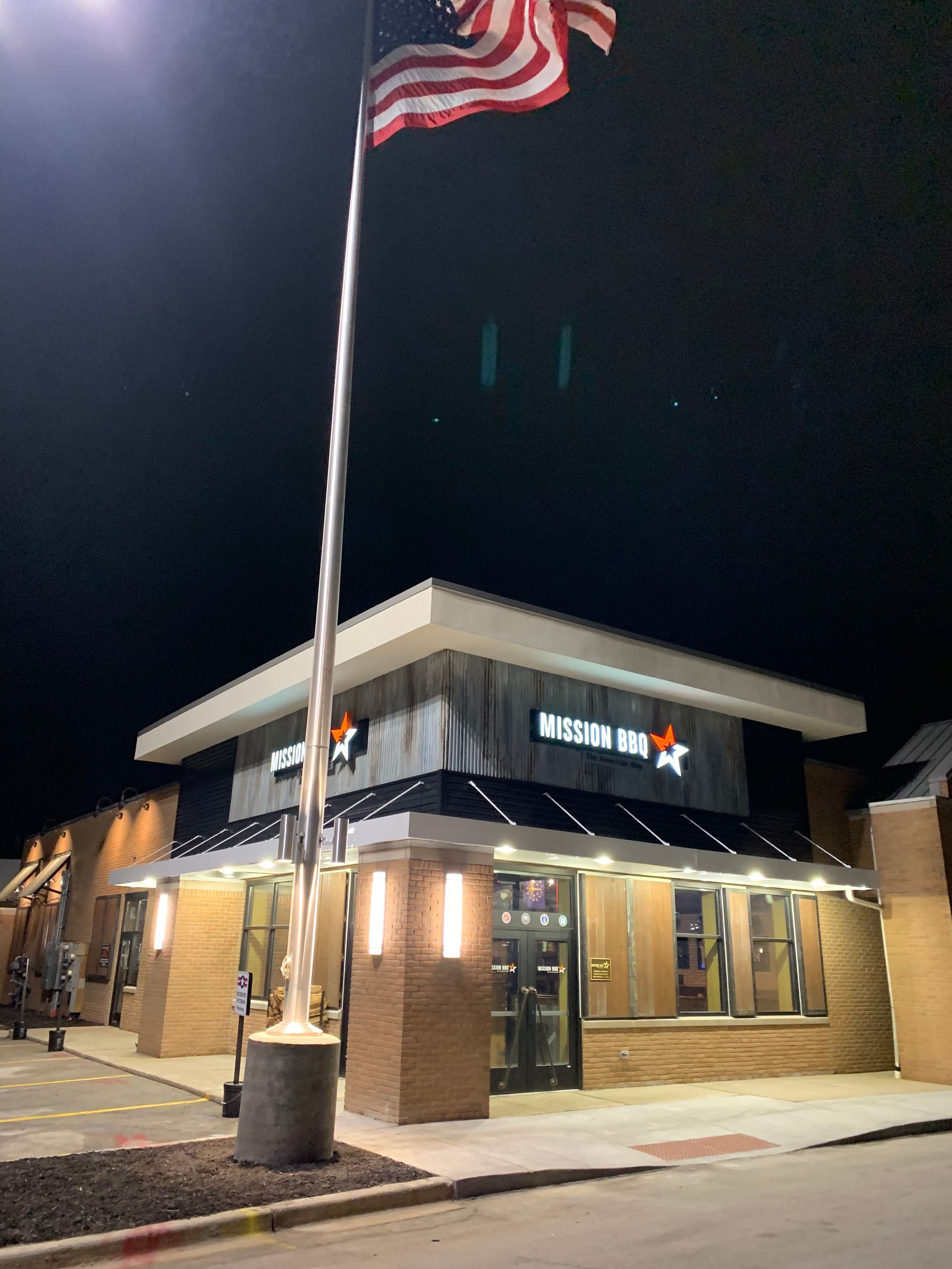 Nighttime exterior of a Mission BBQ restaurant with an American flag and lit entrance.