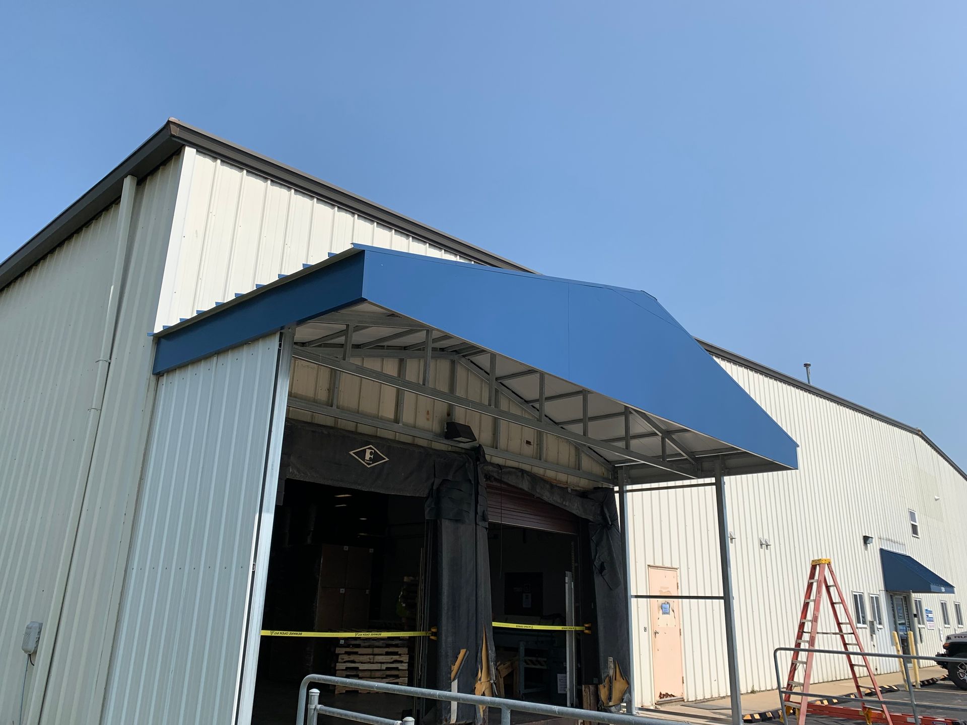 Industrial building with blue awning over an open loading bay and ladder beside it