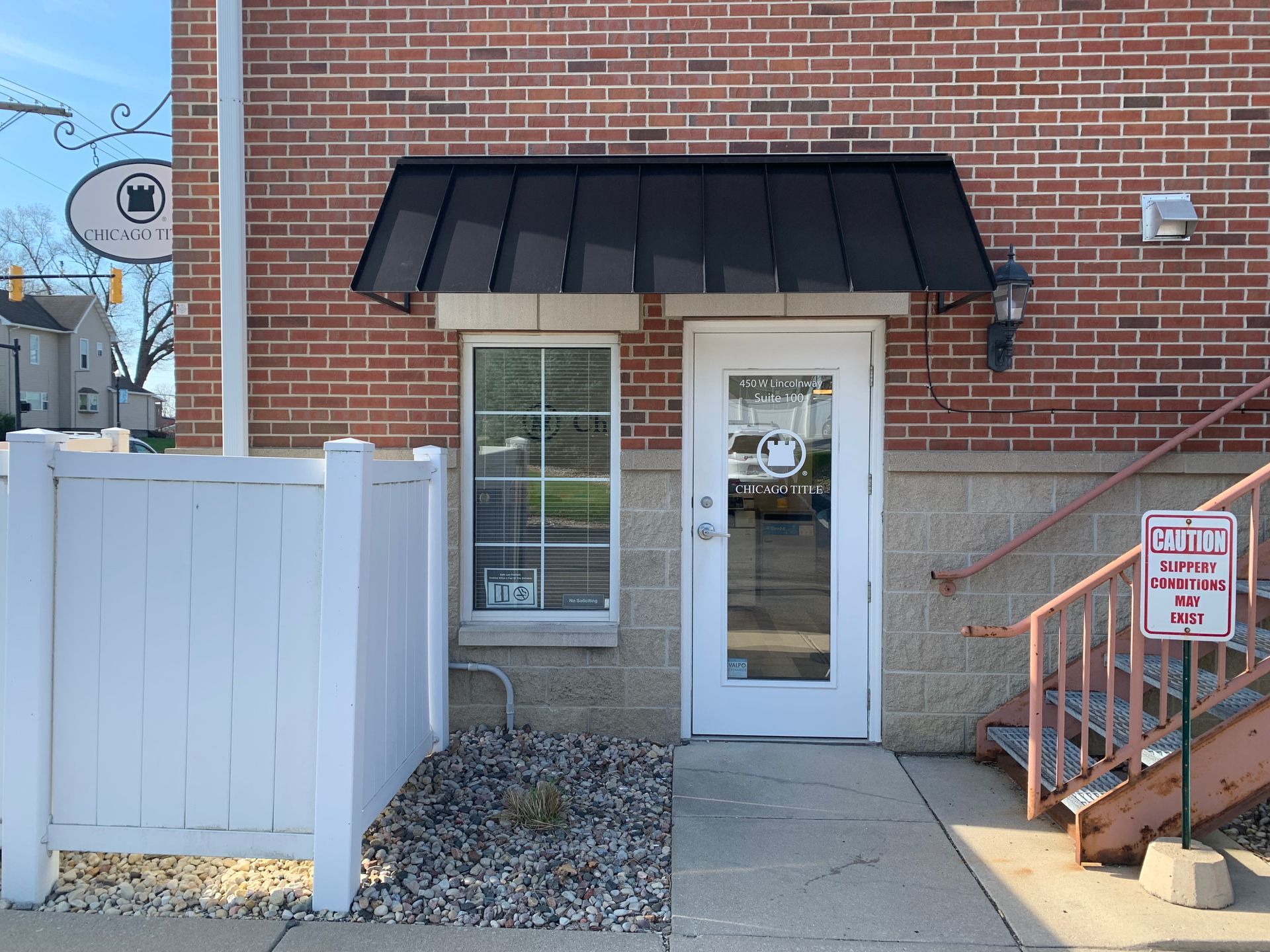 Brick building entrance with white door, black awning, and small stairway beside a fenced walkway.