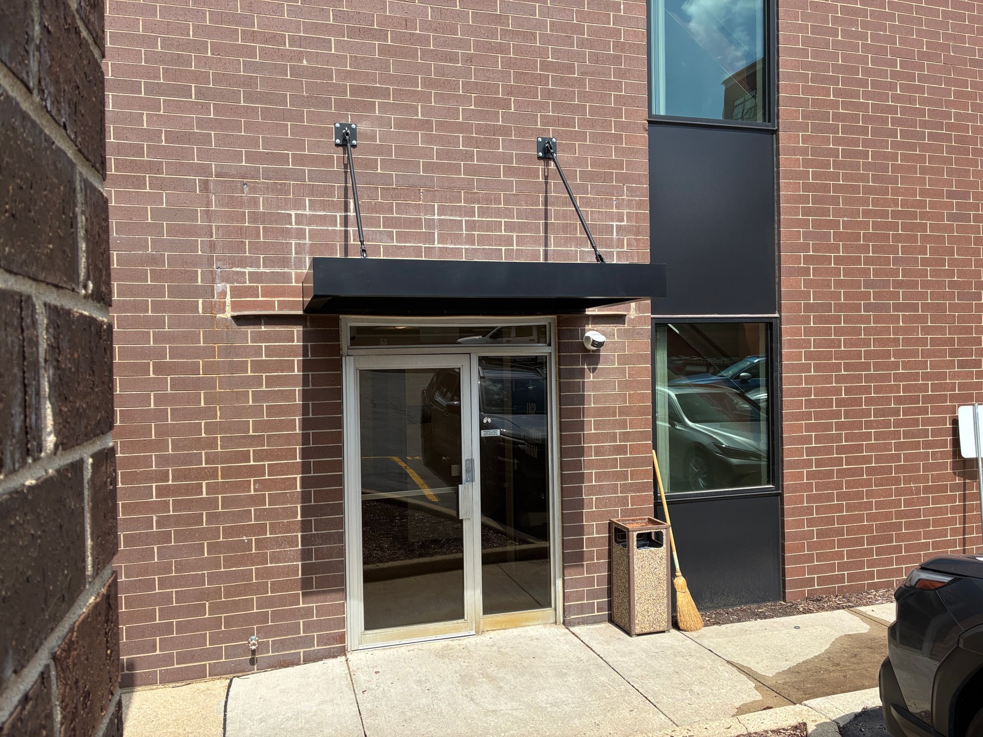 Brick building entrance with glass double doors, black awning, and narrow side windows.