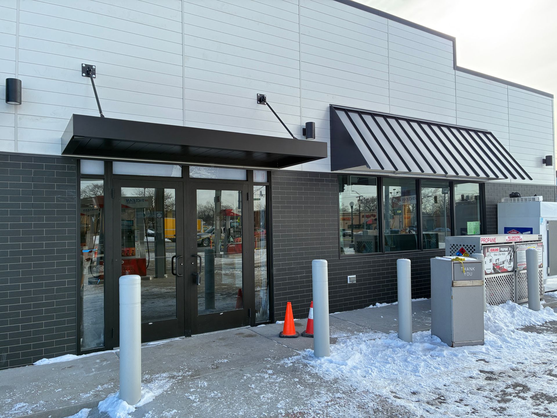 Snowy storefront entrance with glass doors, striped awning, and metal bollards outside