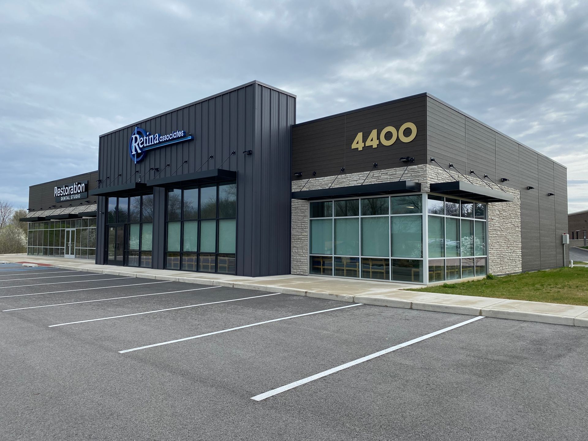 Modern commercial building with glass storefronts, black and stone exterior, and empty parking lot under cloudy sky