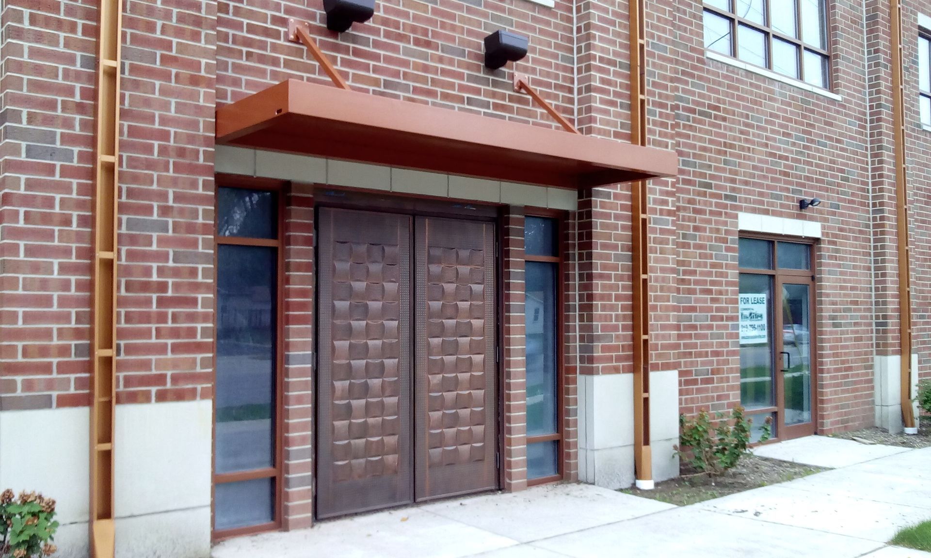 Brick building entrance with double wooden doors, awning, and windows beside a sidewalk.