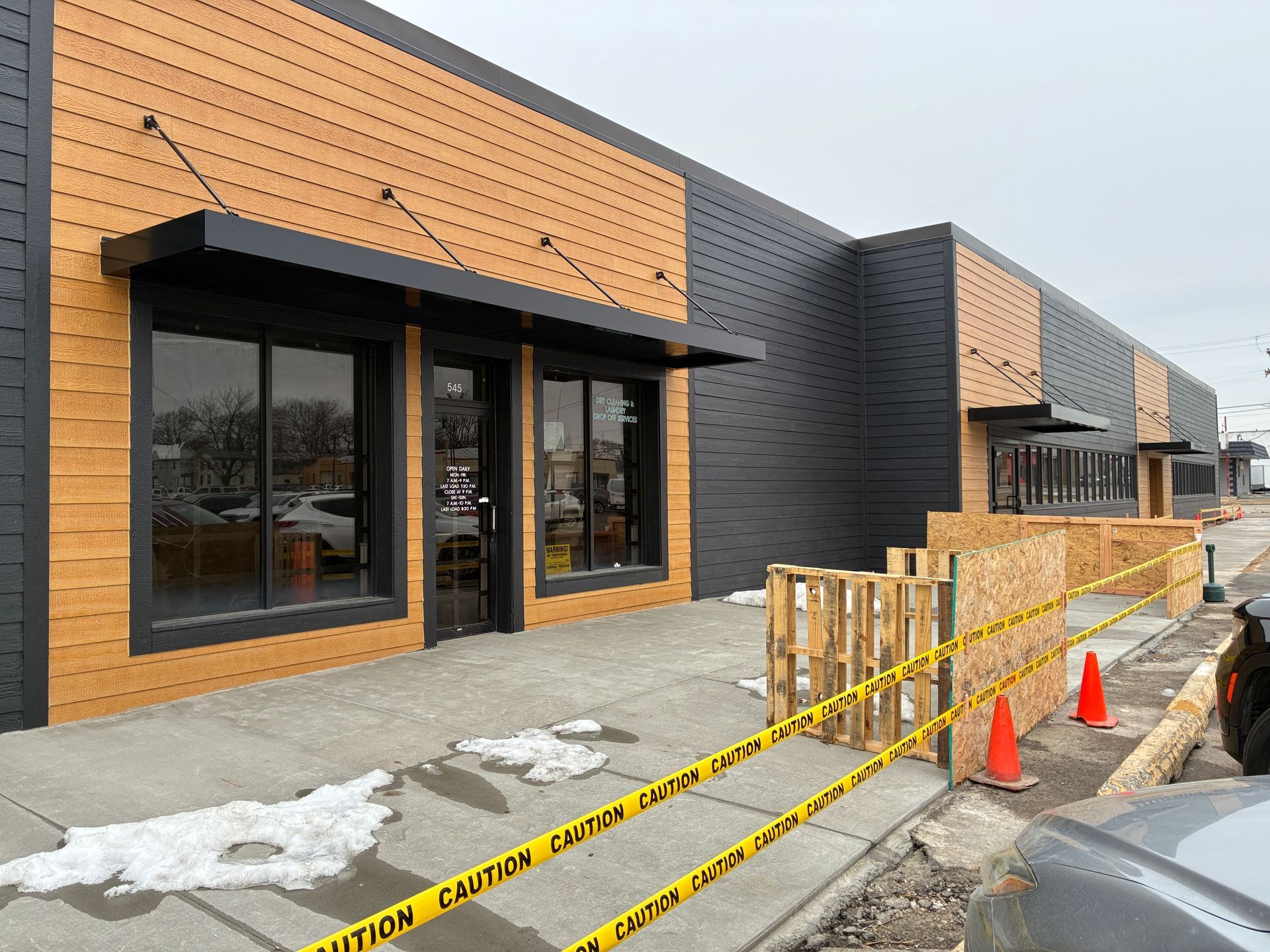 Storefronts under construction with black and wood facades, caution tape, and orange cones along the sidewalk