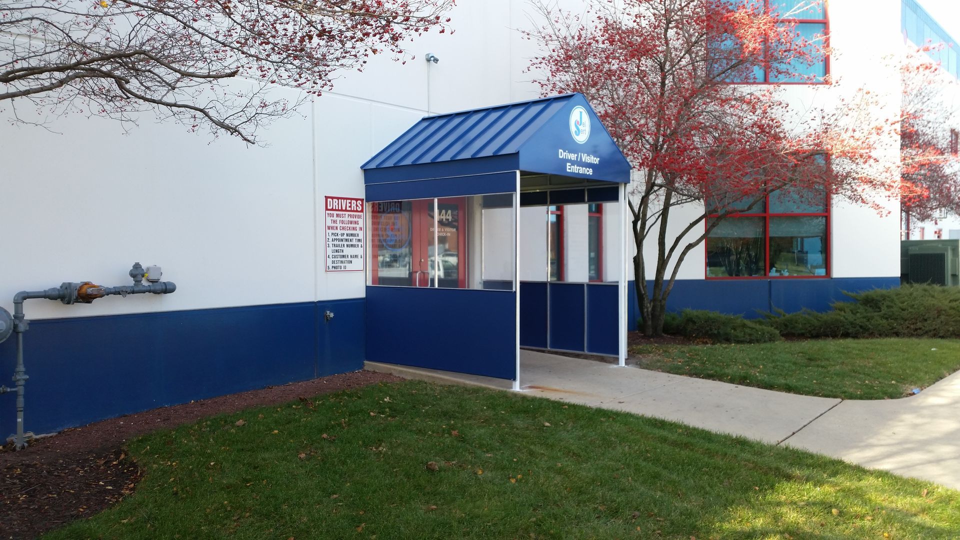 Blue awning entrance to a building with a sidewalk, lawn, and red-leaf trees.
