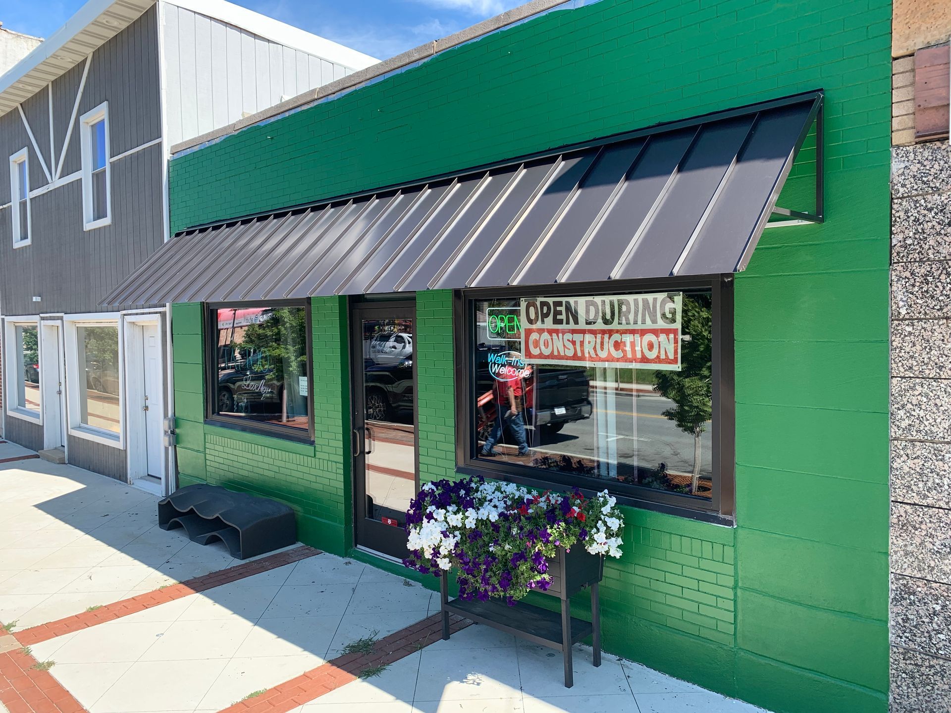 Green storefront with black awning and sign reading “Open” and “Construction,” plus flowers by the window.