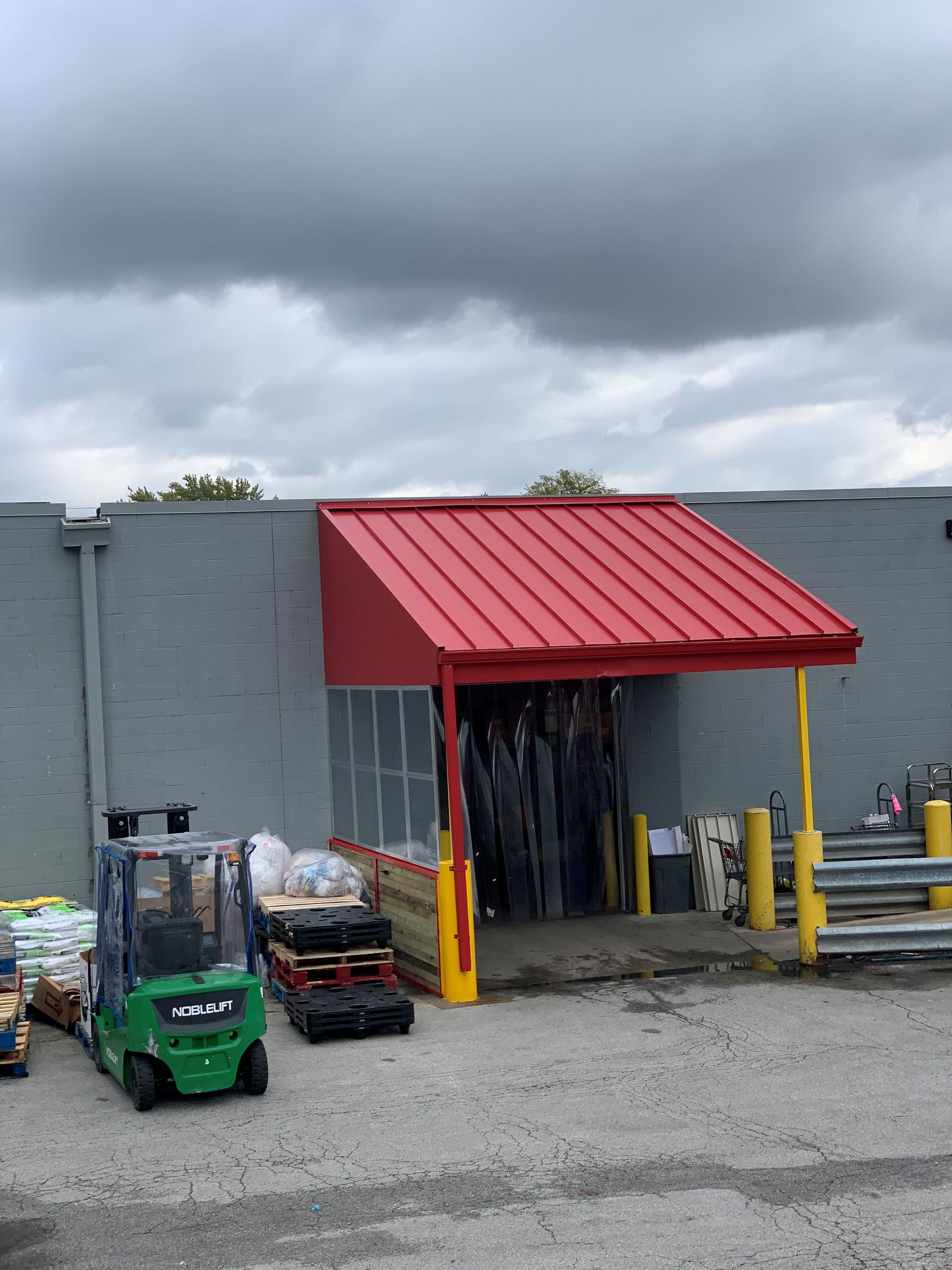 Outdoor storage area with gray wall, red awning, stacked items, and carts on gravel under cloudy sky
