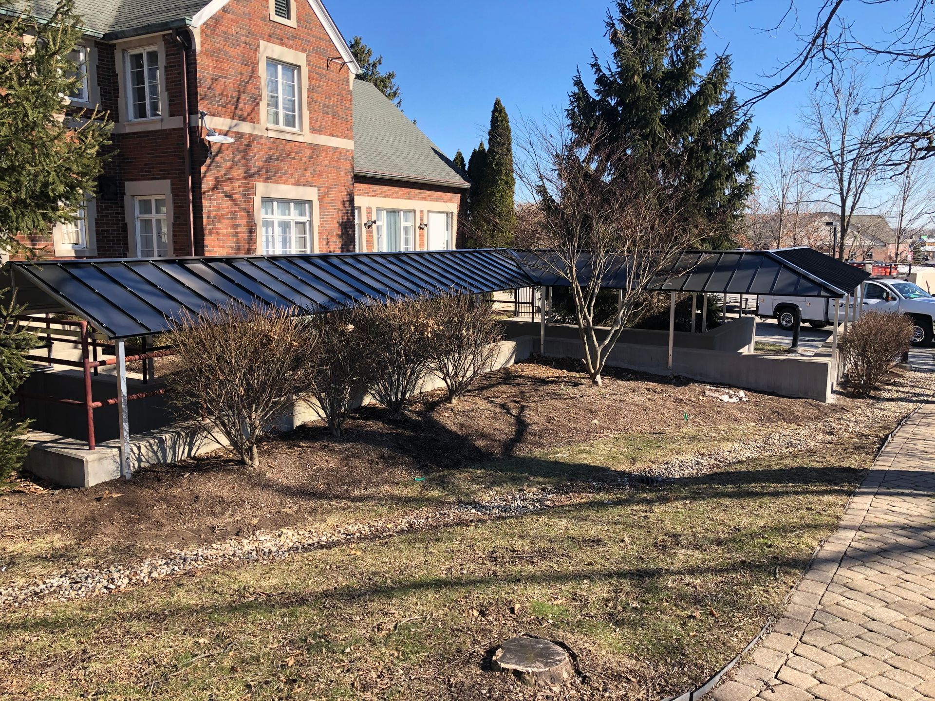 Brick building with a sloped blue roof, leafless shrubs, and a paved walkway in a sunny courtyard
