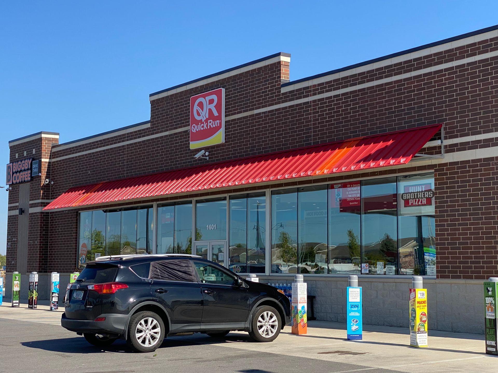 Grocery store with red awnings and parked black SUV under a clear blue sky