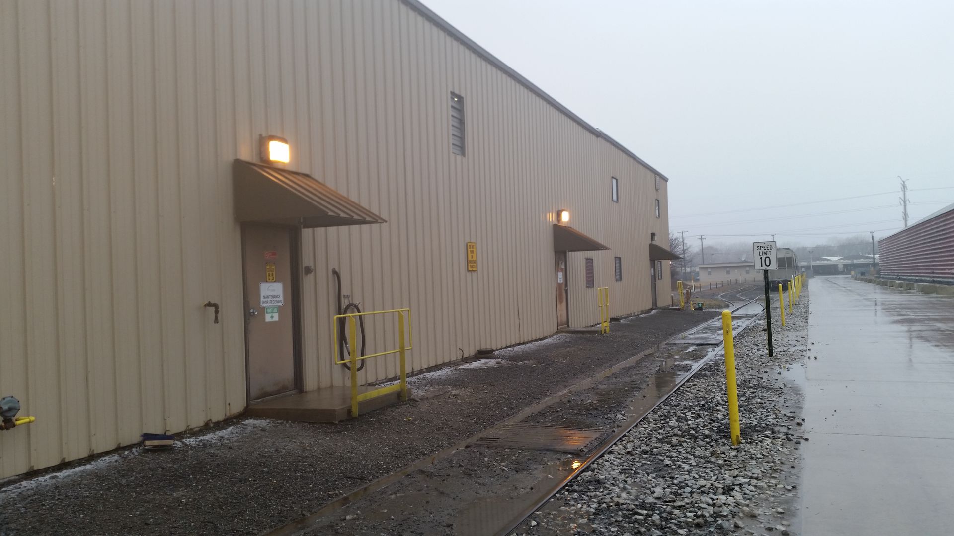 Foggy industrial loading dock beside a beige warehouse, with wet gravel and a flooded roadway.