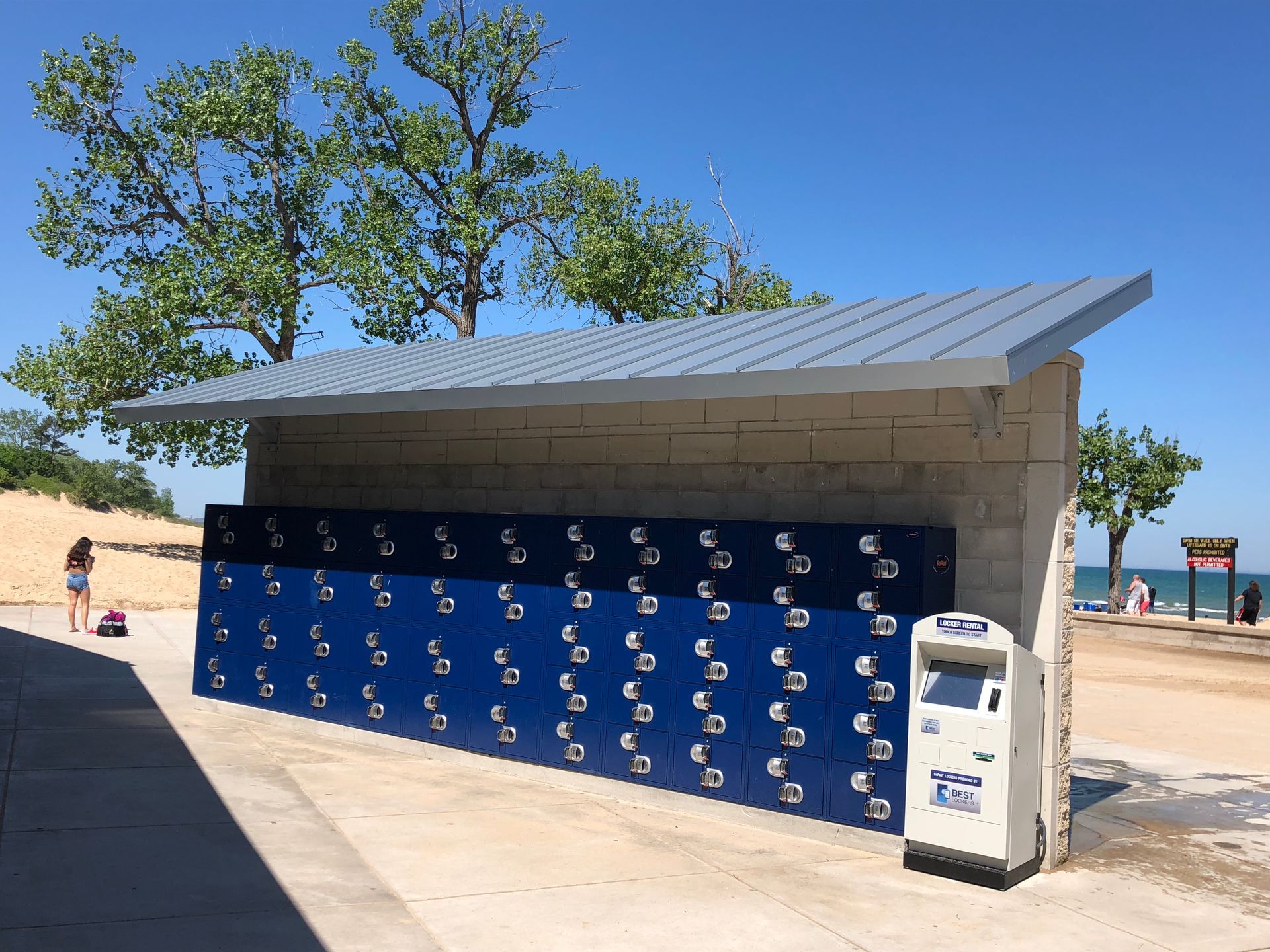 Blue beachside locker wall under a shaded shelter with trees and sand nearby