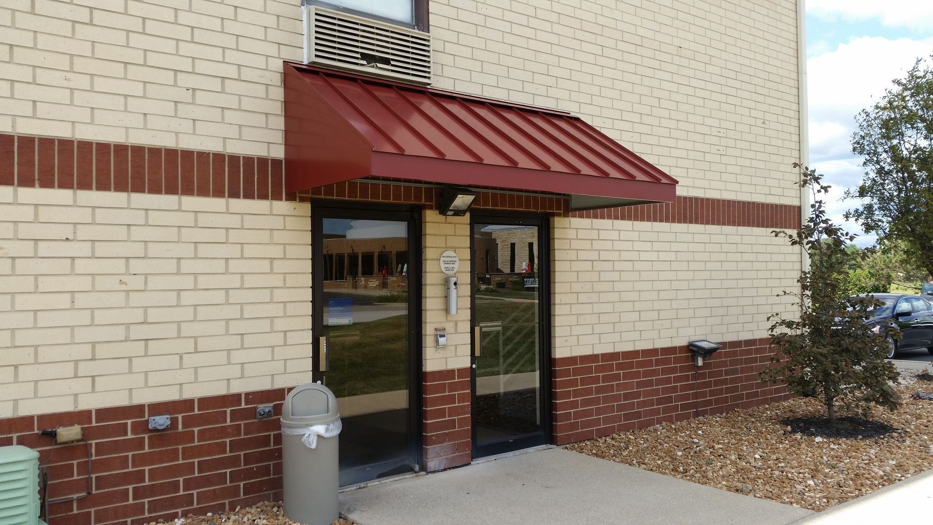 Building entrance with red awning, glass door, brick trim, and a trash can outside