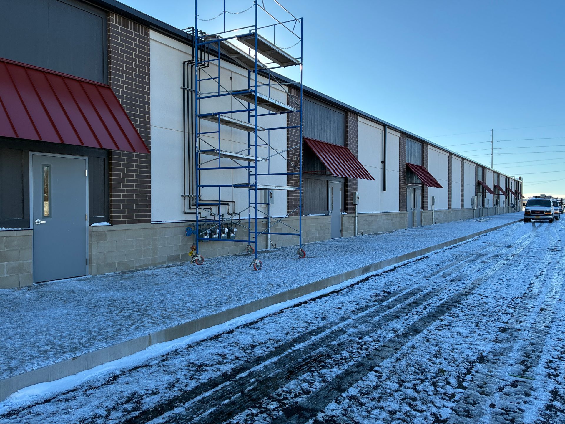Snowy industrial building with red awnings and a scaffolding tower beside a gravel road