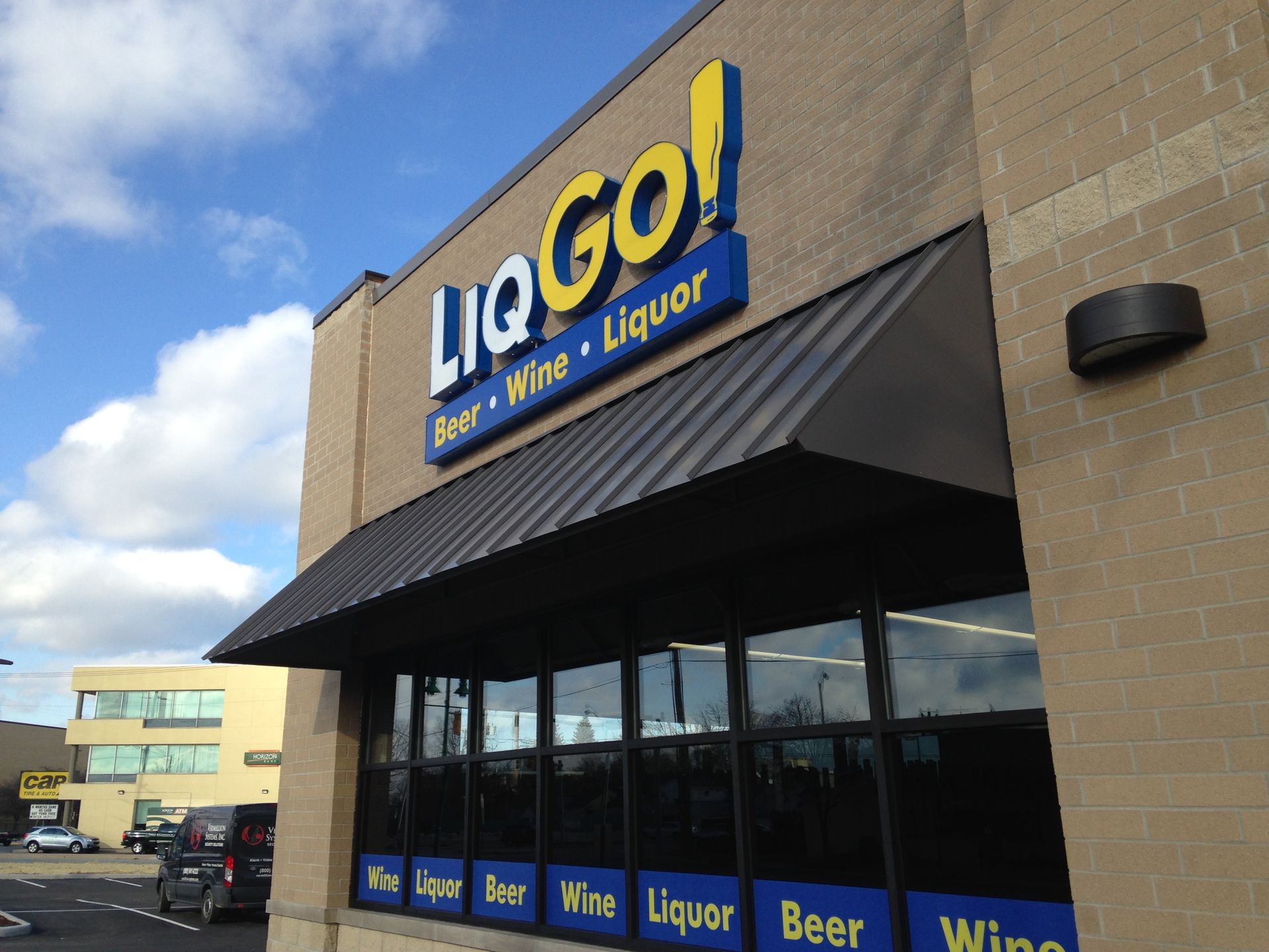 Liquor store storefront with black awning and blue-yellow “Lqgo!” sign under a cloudy sky