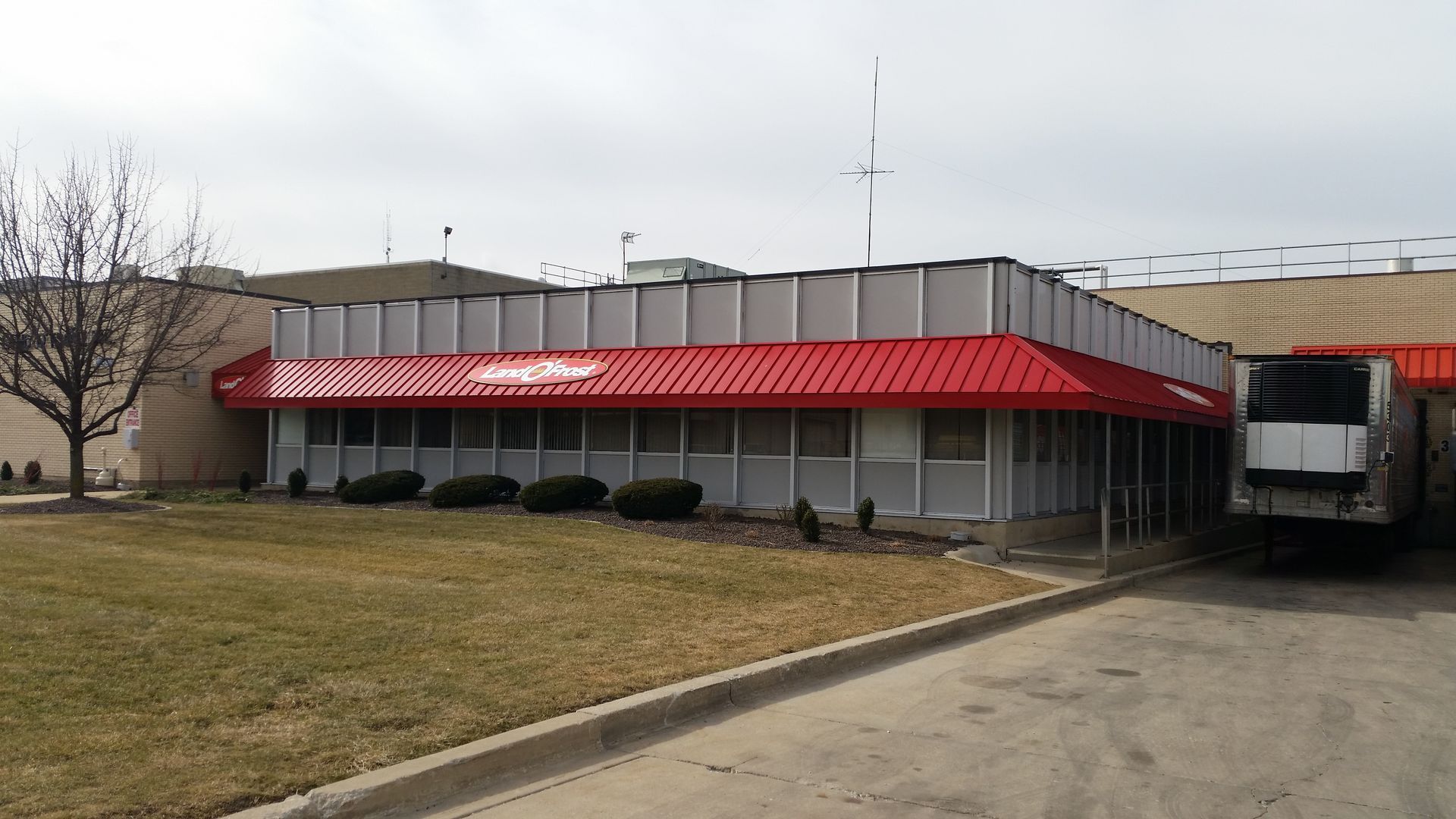 Low beige building with red awning and truck dock, viewed from a grassy roadside.