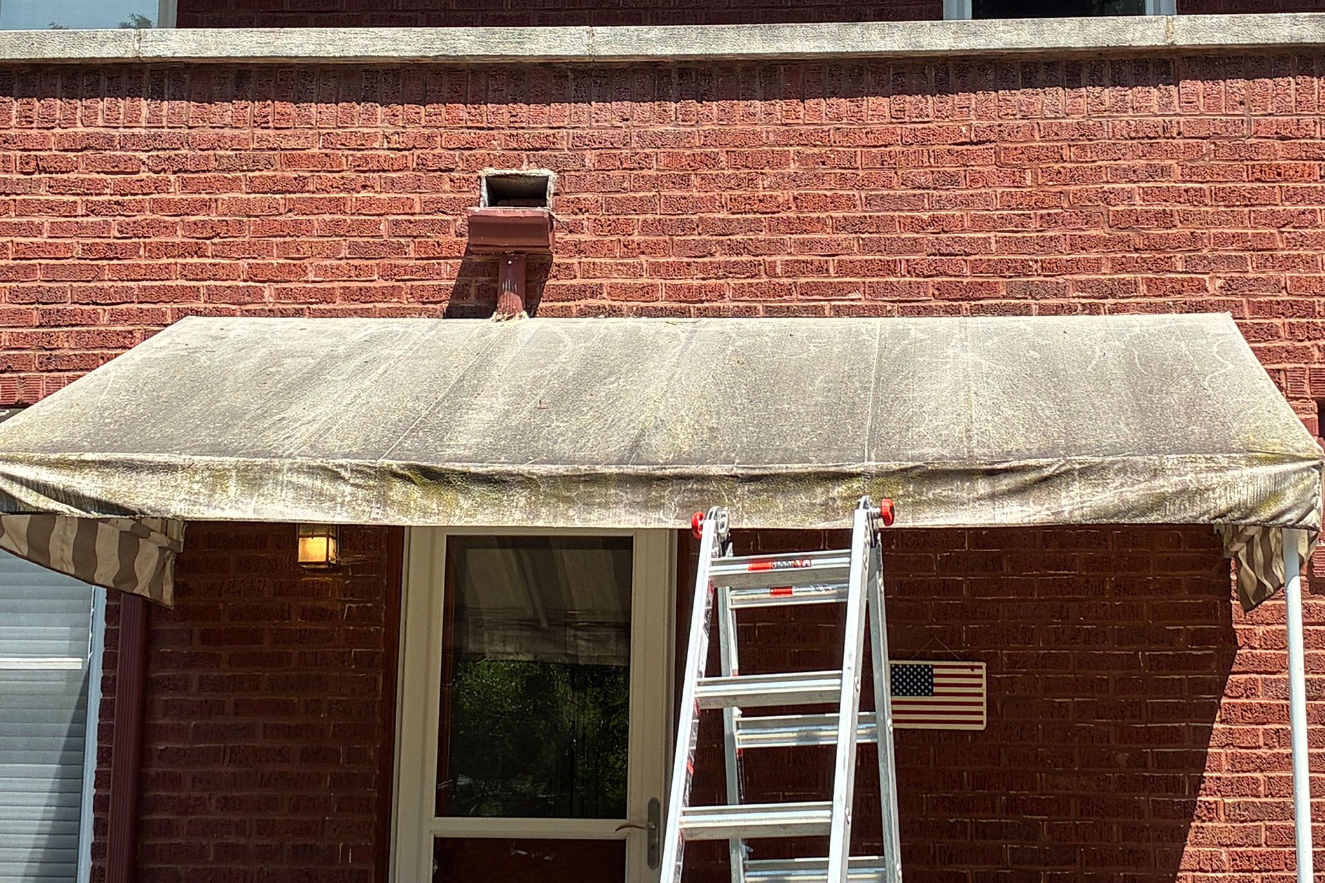 Ladder on a brick house porch beneath a beige awning, with an American flag by the door