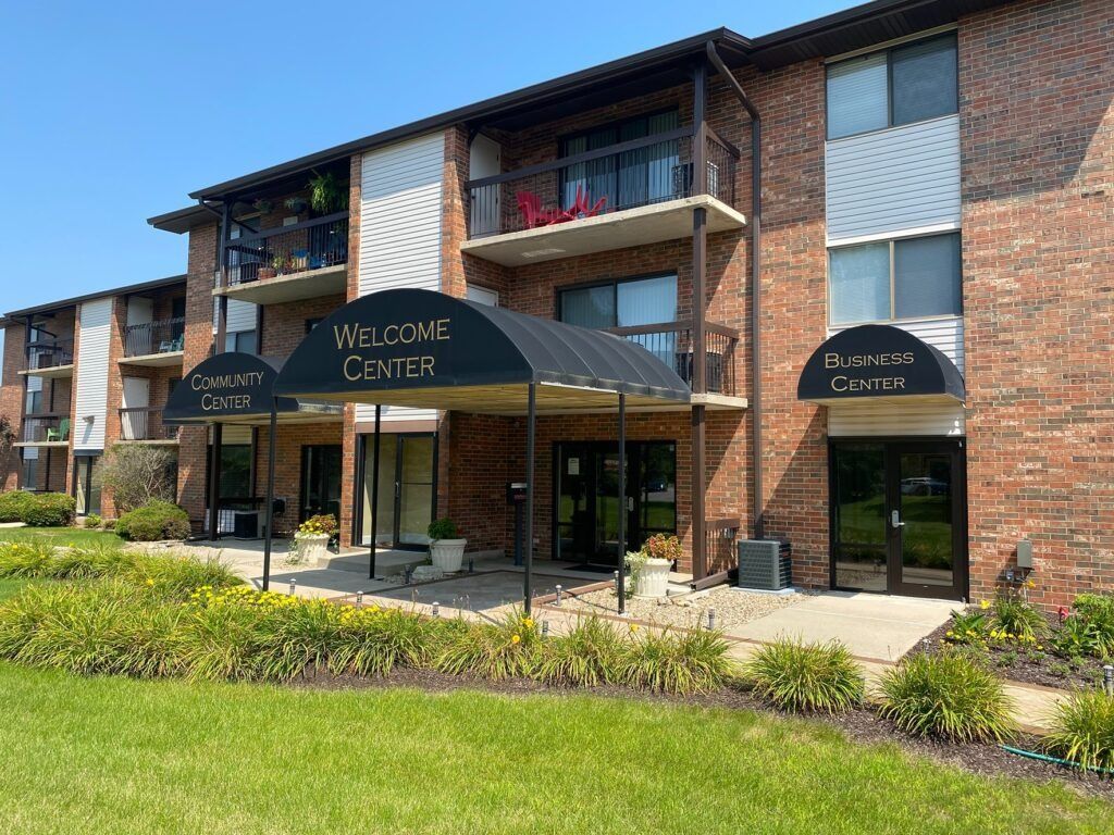 Apartment building entrance with black awnings and a welcome canopy, fronted by a grassy lawn and shrubs