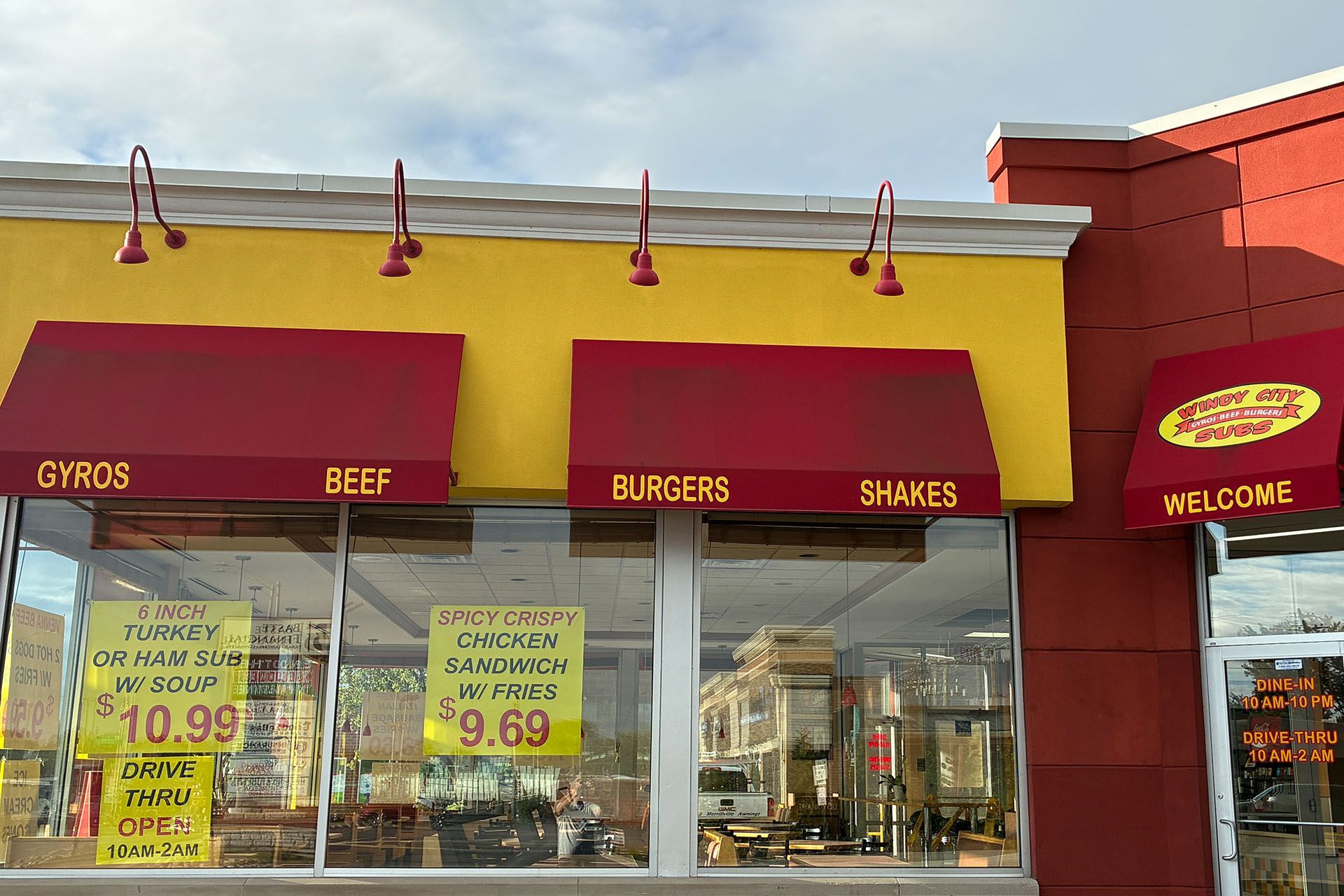 Fast-food restaurant exterior with red awning, yellow signs, and menu posters for gyros, beef, burgers, and shakes