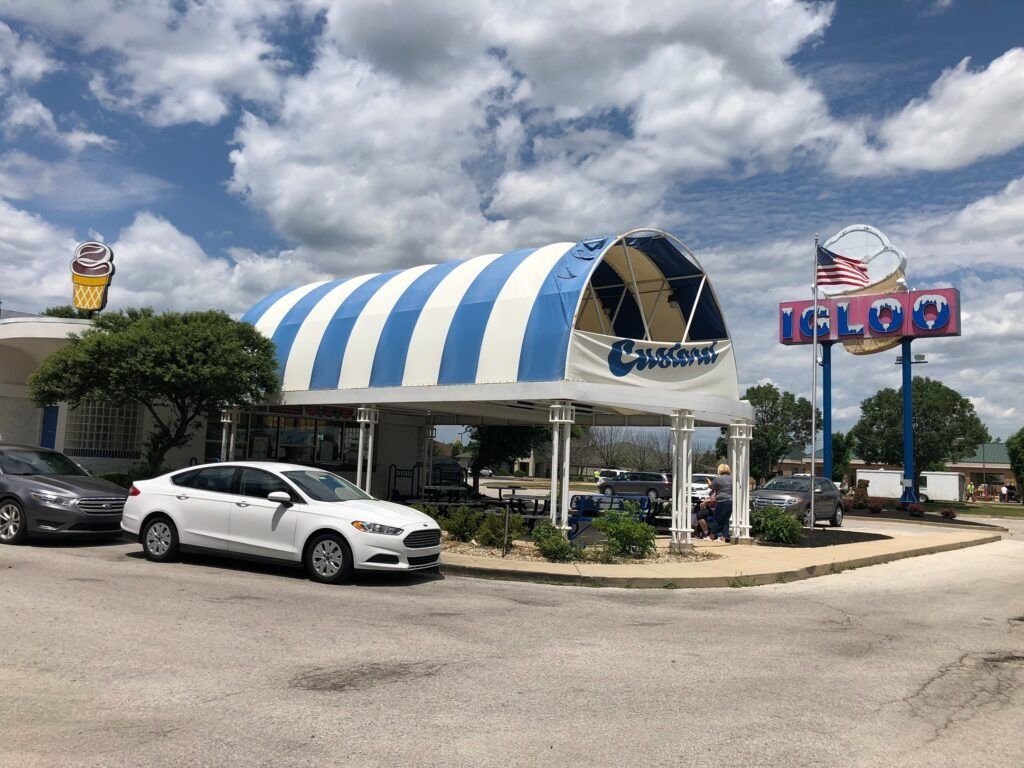 Blue-and-white striped drive-in restaurant with parked cars under a cloudy sky