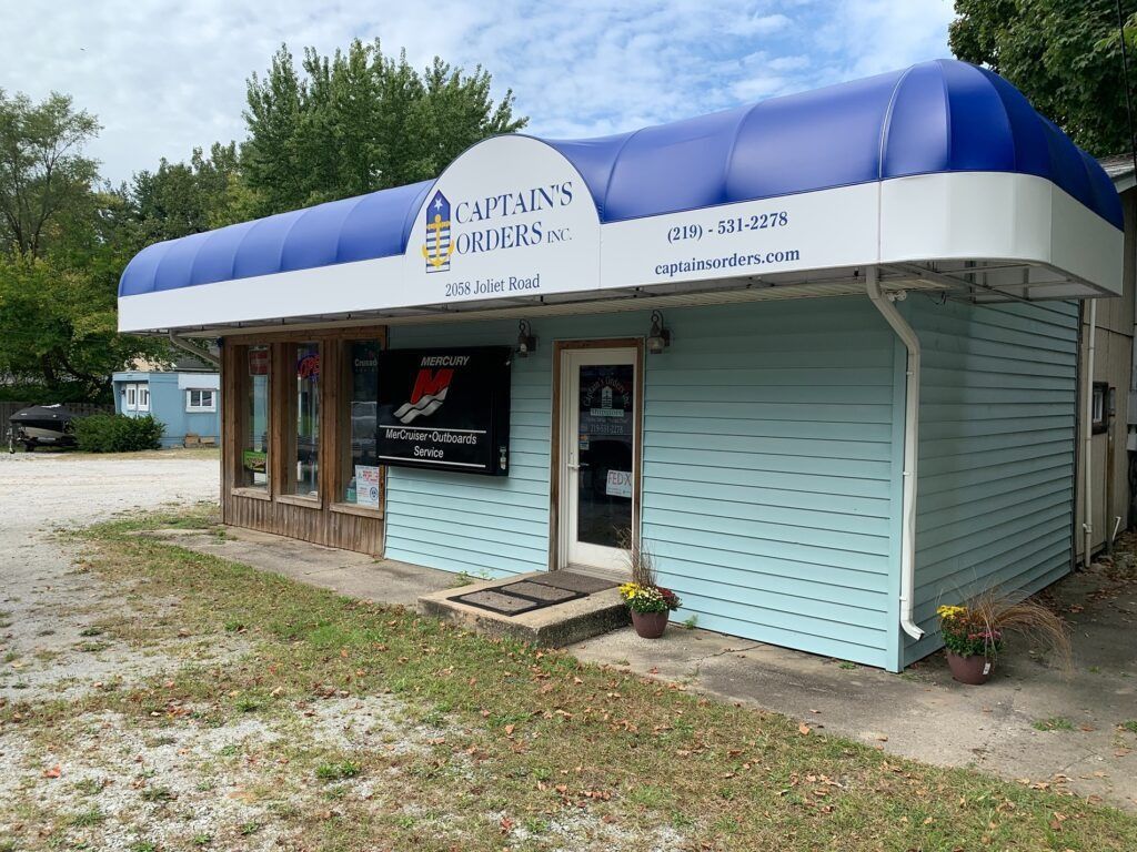 Small storefront with blue-and-white awning and siding, set beside a gravel lot and trees.