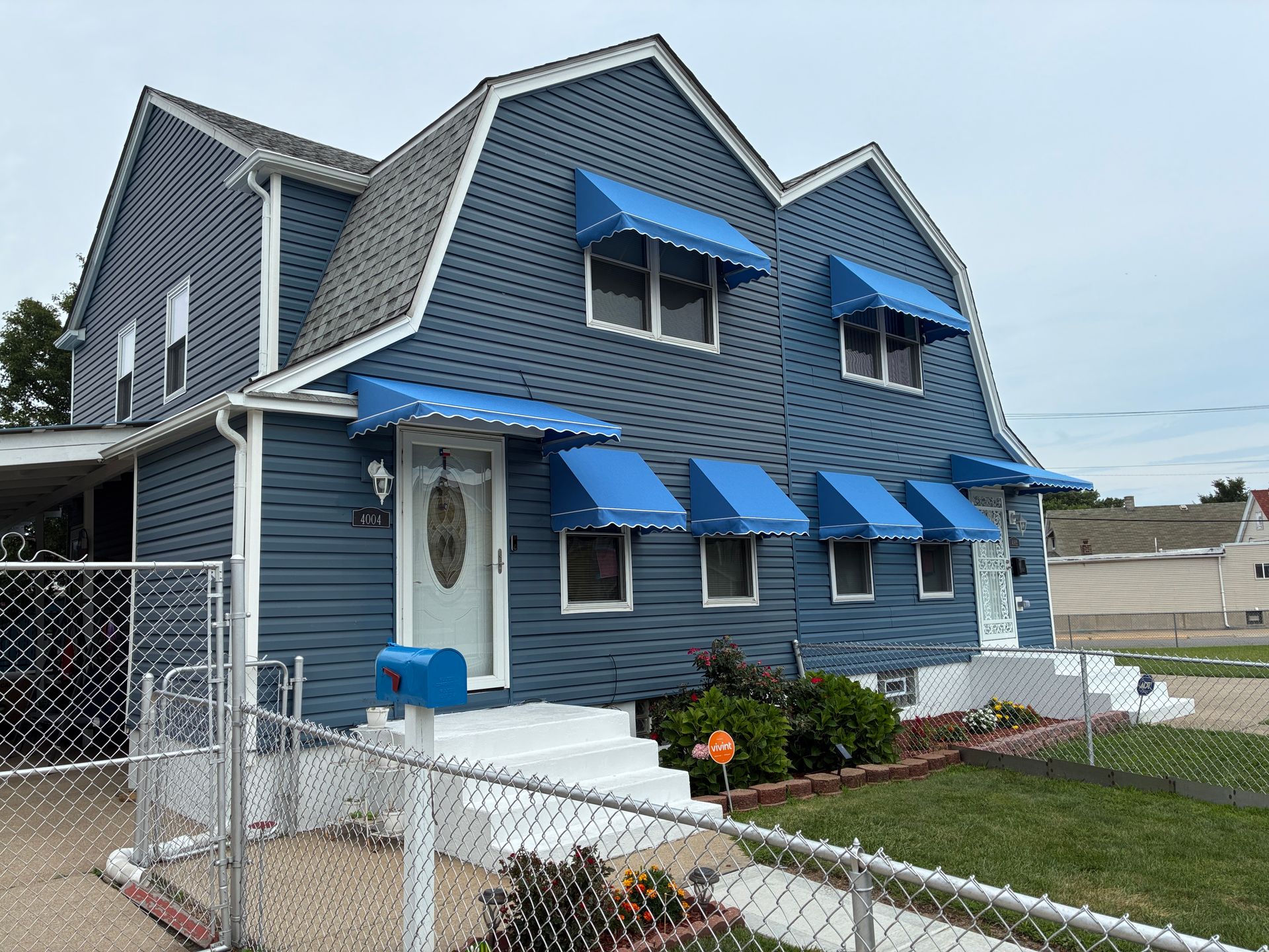 Blue two-story house with white trim and blue awnings beside a fenced yard