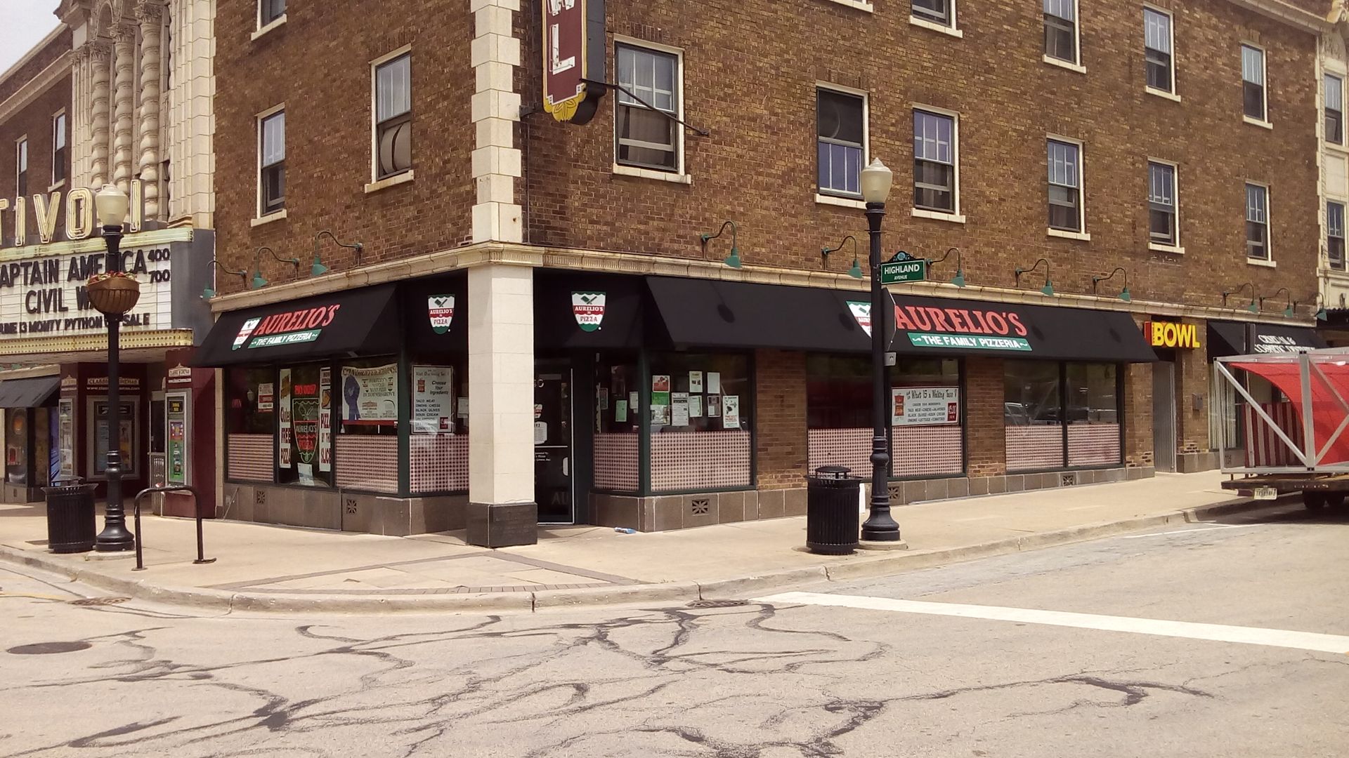 Street corner with a brick building, storefronts, and a bus stop under an overcast sky