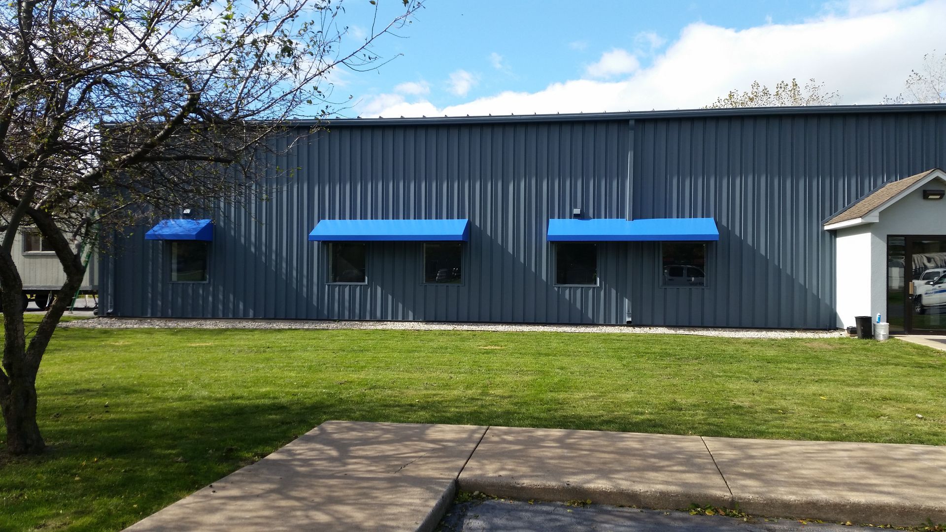 Gray metal building with bright blue awnings and a small tree in a grassy yard