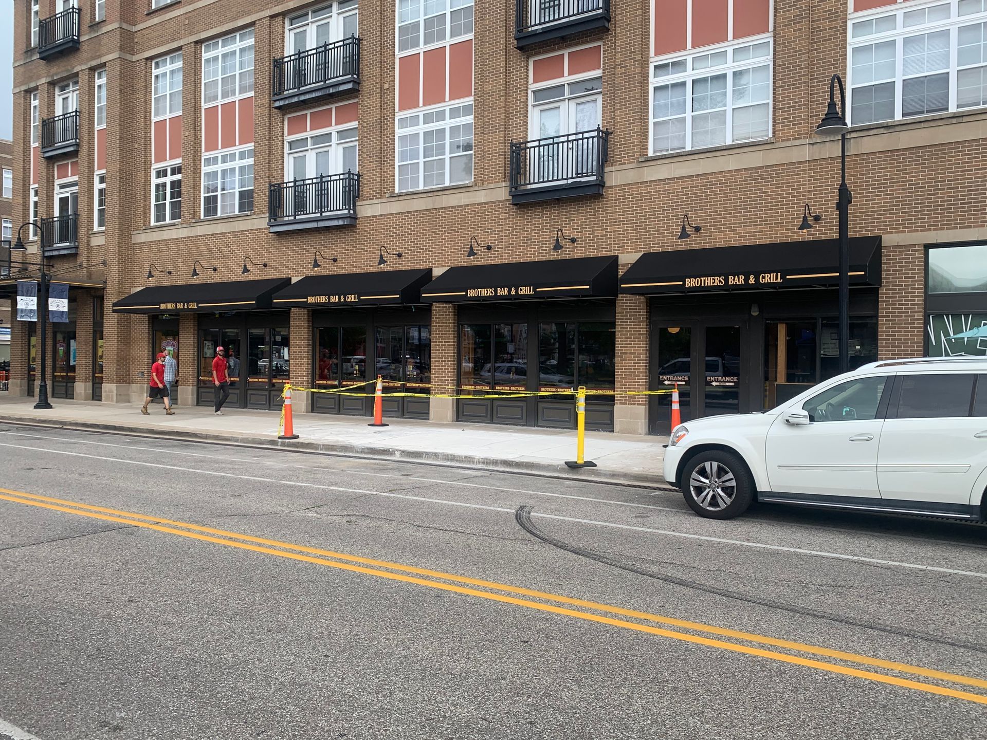 Brick storefront with awnings and parked white SUV on a city street, pedestrians walking by.
