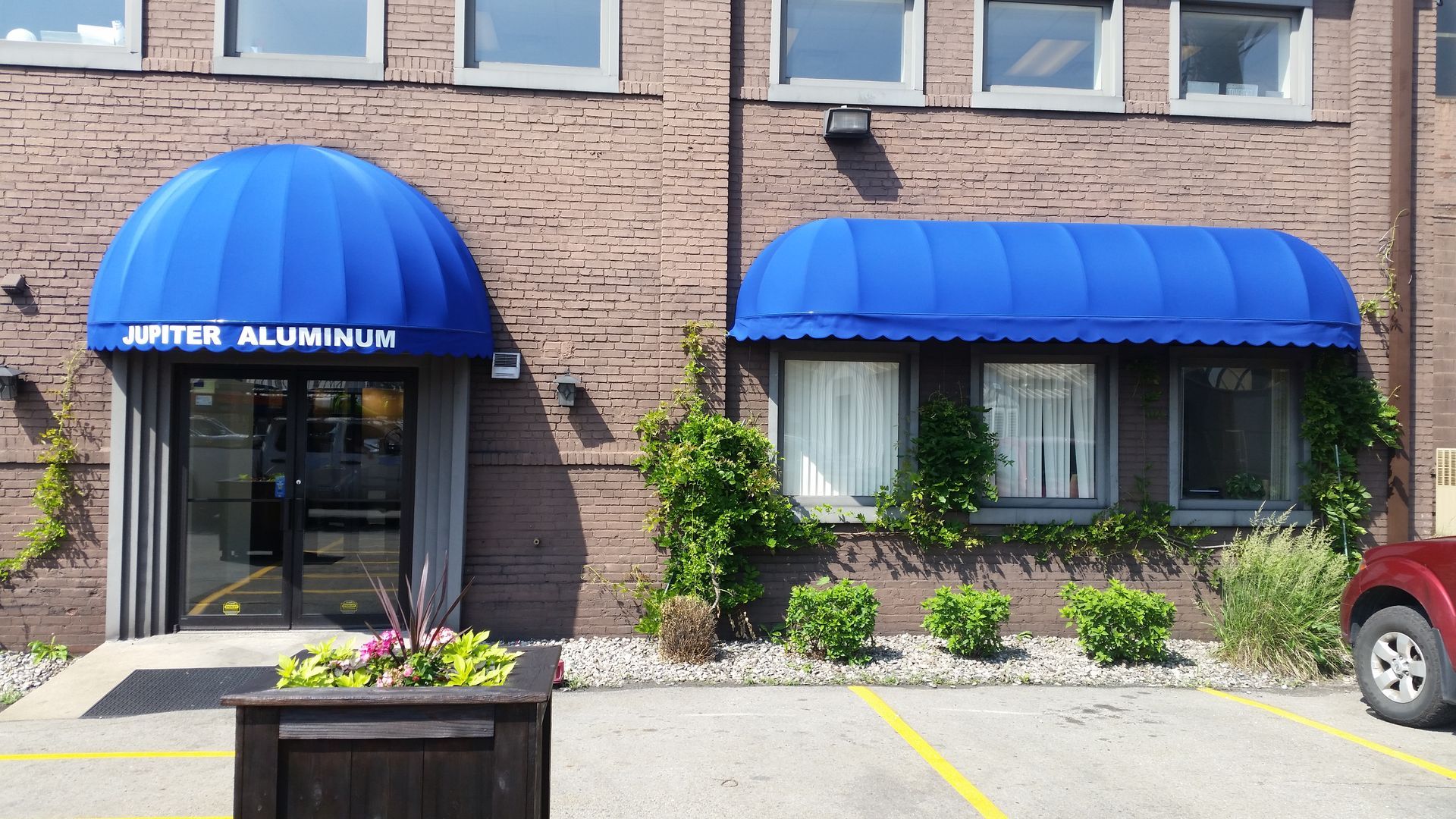 Brick storefront with blue awnings, glass entrance, and flower planter in front of parking spaces