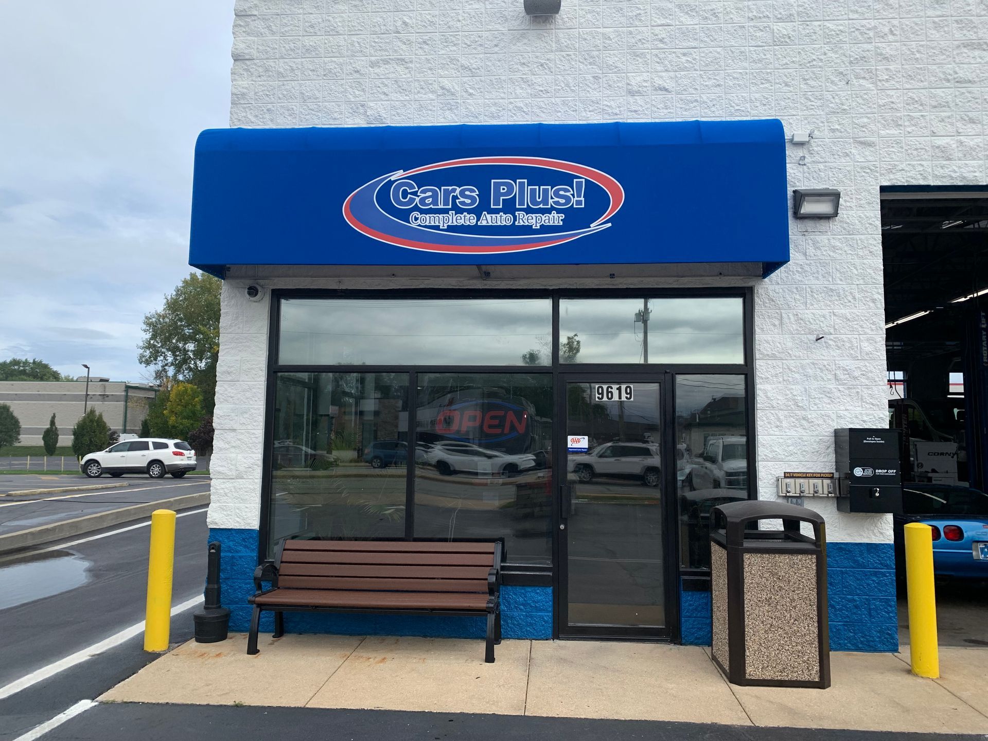 Cars Plus convenience store entrance with blue awning, bench, trash can, and yellow bollards