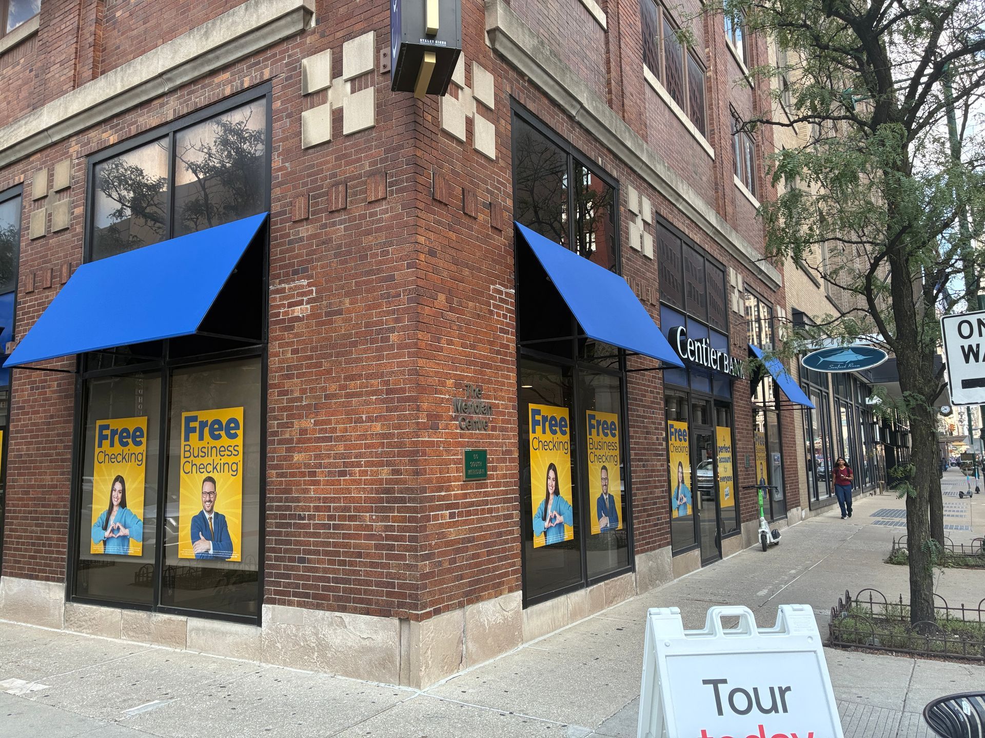 Corner storefront with blue awnings, brick facade, and a sidewalk sign reading “Tour.”