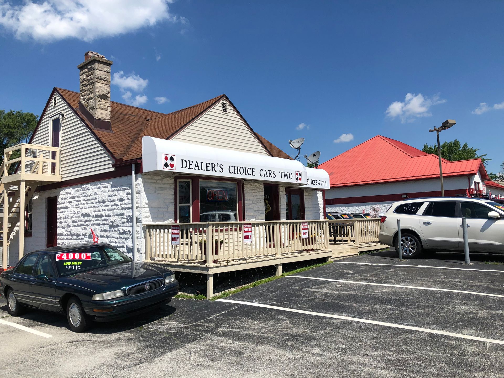 Small white storefront with porch, parked cars, and a red-roofed building under a blue sky
