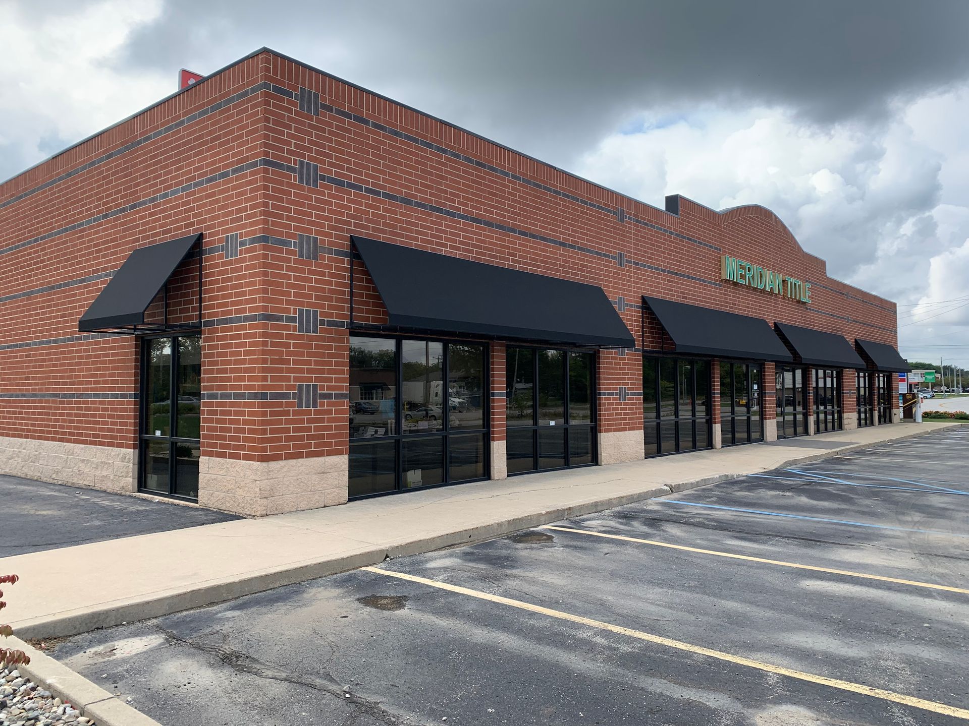 Brick storefront with black awnings and green sign in an empty parking lot under cloudy skies