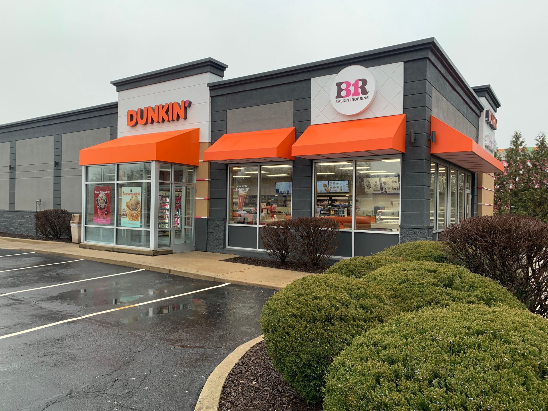 Dunkin’ storefront with orange awnings and a BB sign on a rainy day, with trimmed bushes in front