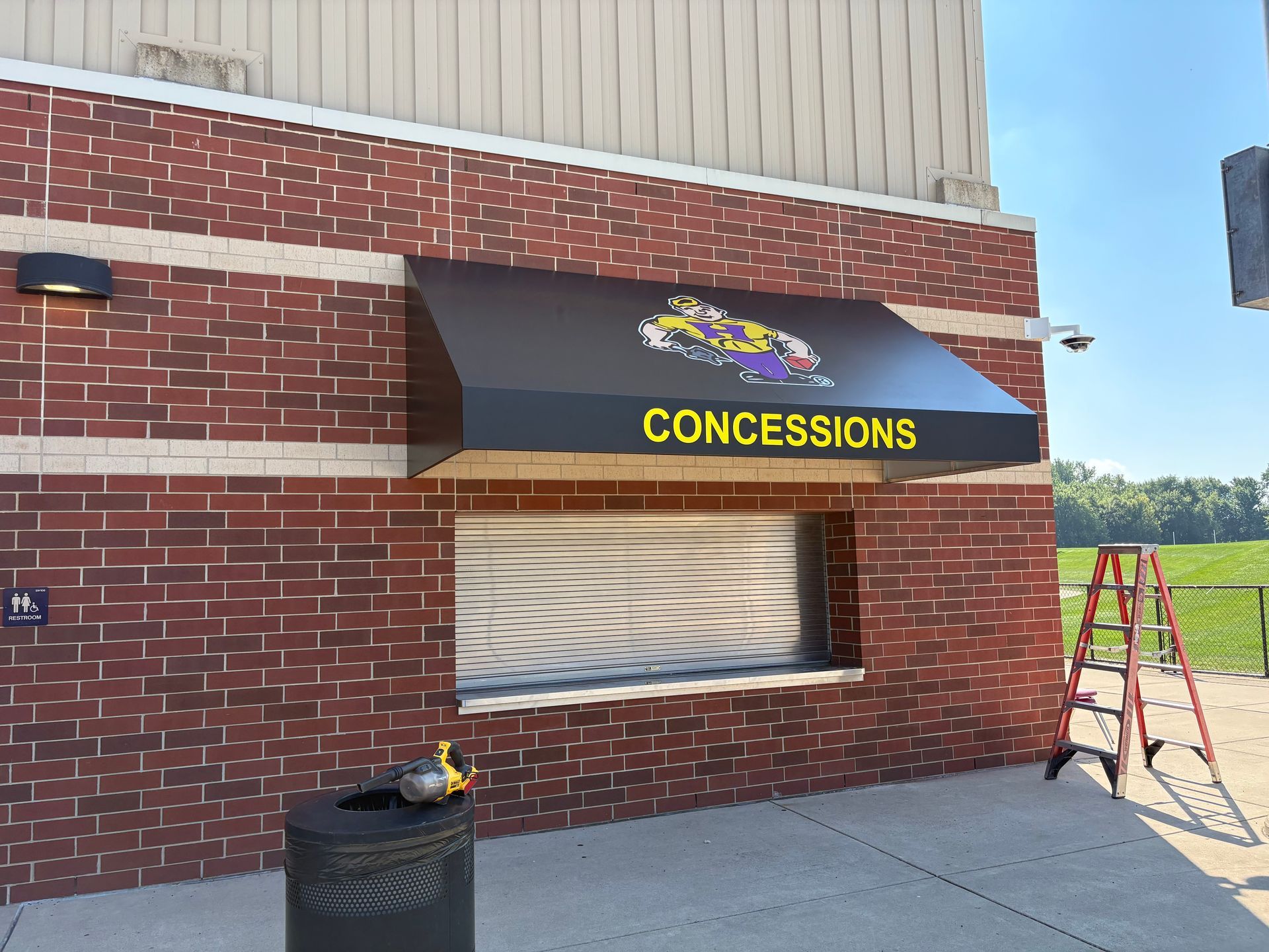 Closed concession stand window on a brick building with black awning and yellow “CONCESSIONS” sign; ladder nearby