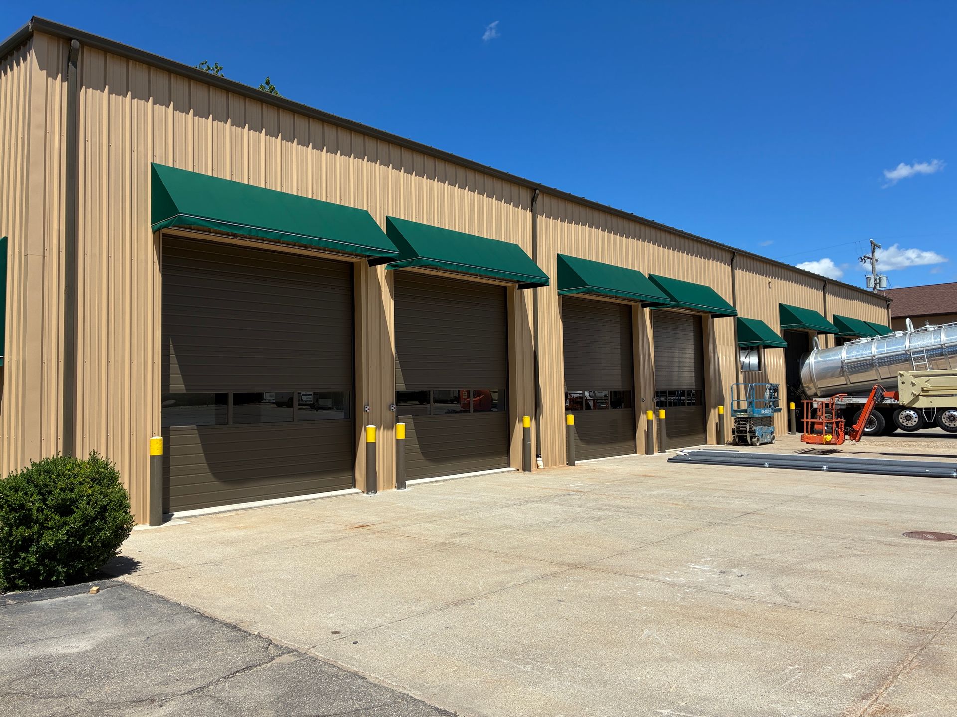 Industrial warehouse loading bays with green awnings and a tanker truck parked beside them under a clear blue sky