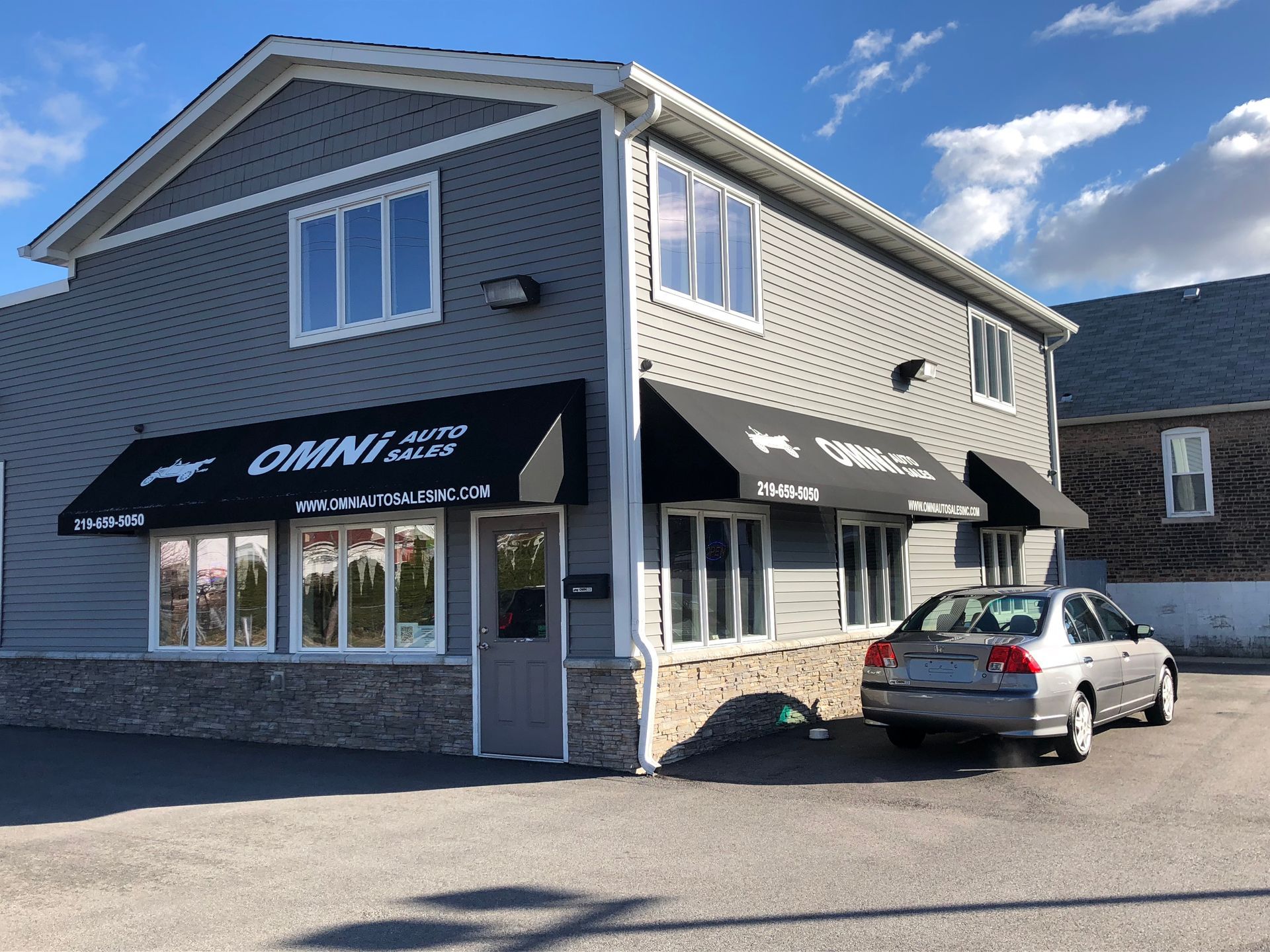 Corner storefront with black awnings, stone base, and a silver car parked outside under a blue sky.