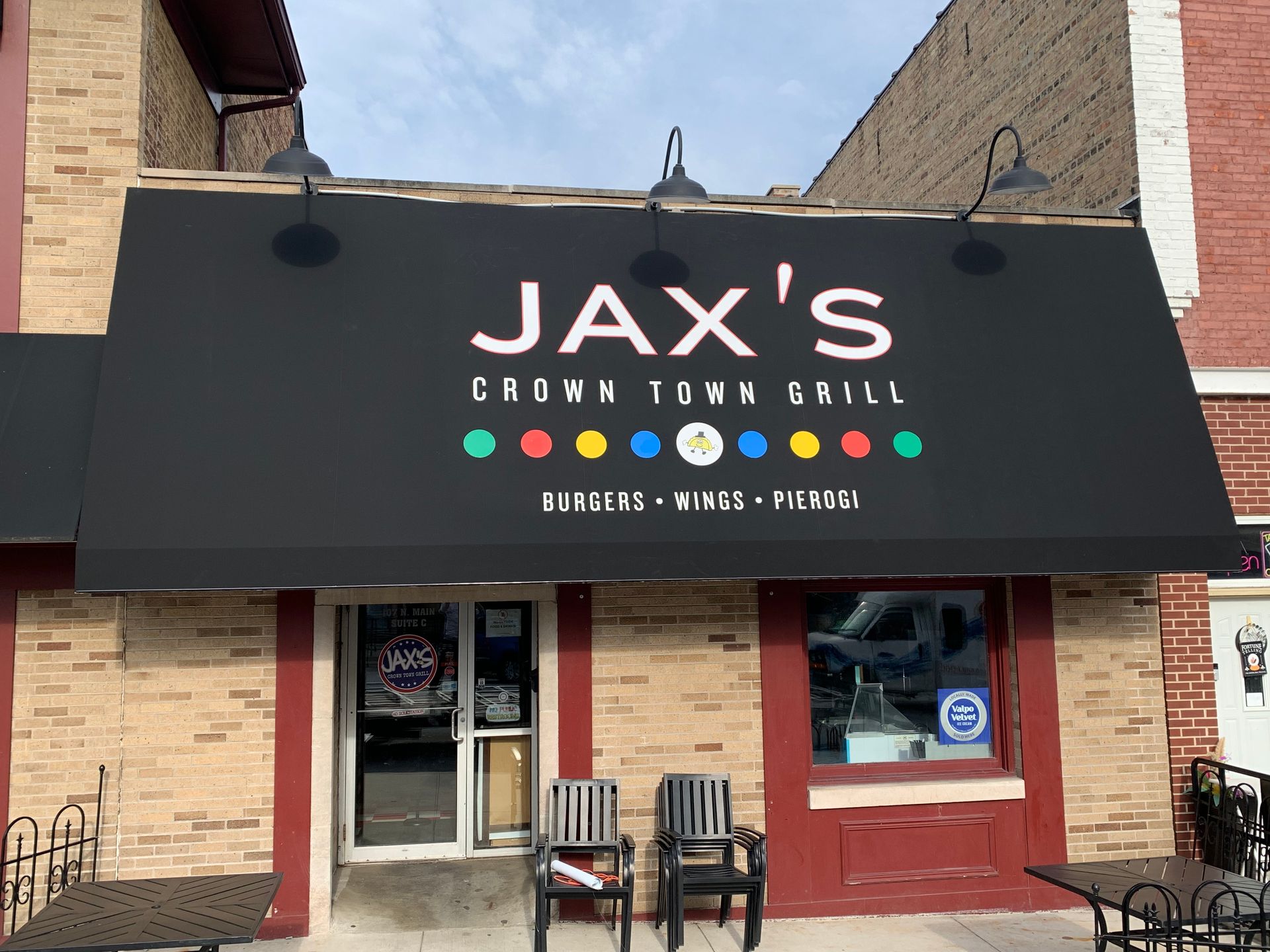 Storefront with black awning reading “JAX’S,” brick facade, and chairs outside